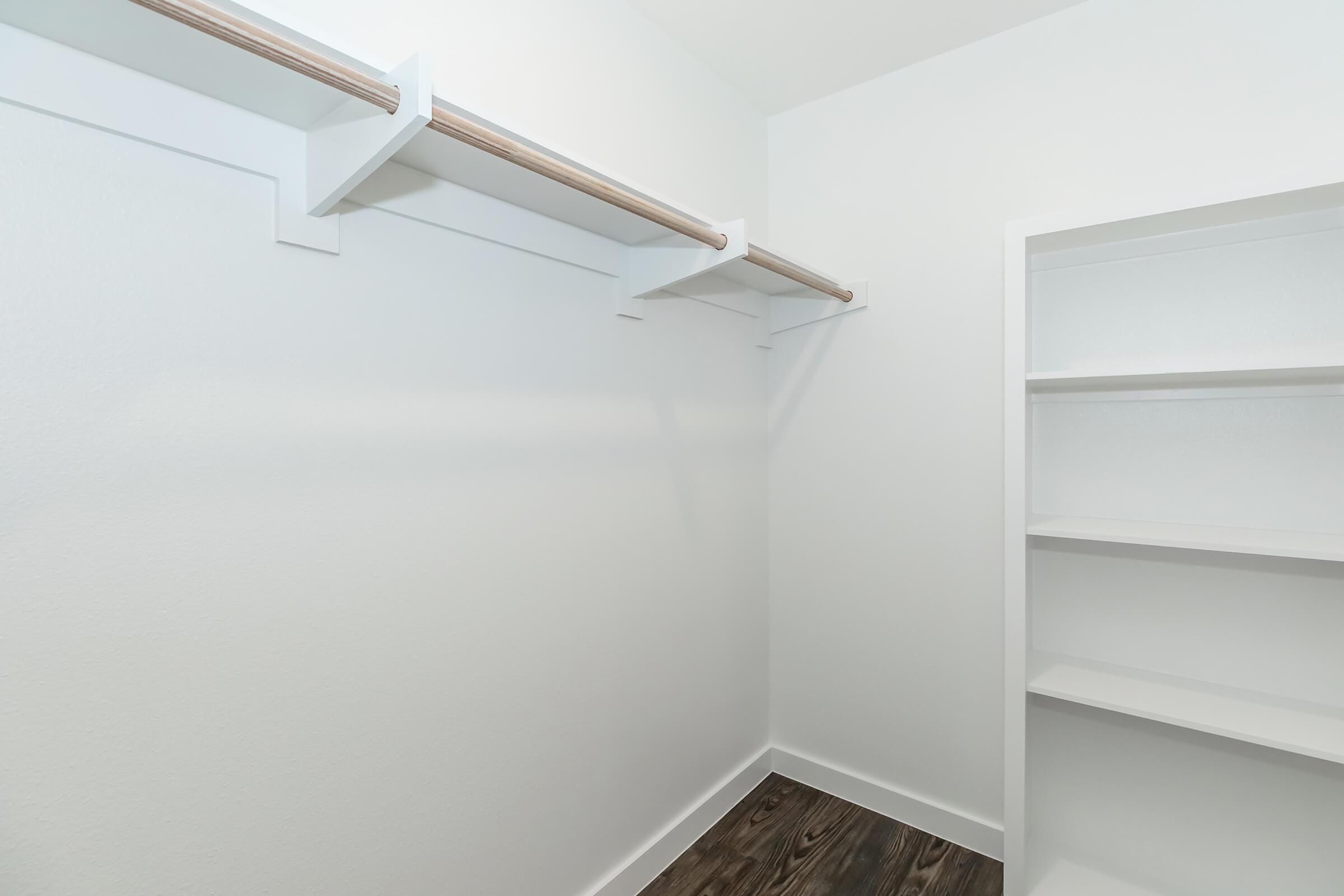 A clean, empty closet featuring a white wall and a wooden rod for hanging clothes. On one side, there is a simple white shelf unit, while the flooring has a dark, wooden finish. The overall ambiance is bright and spacious, emphasizing minimalism and organization.