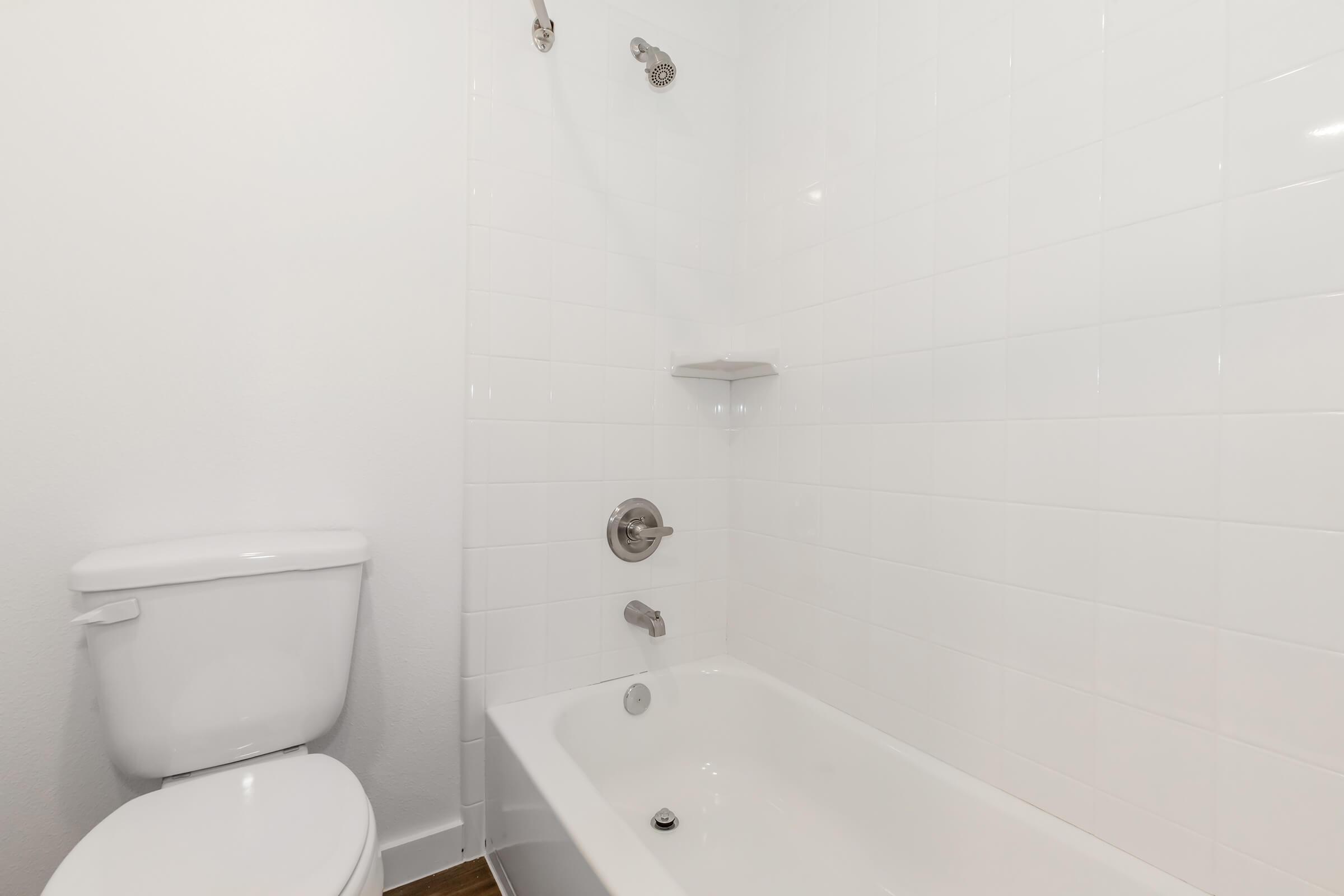 A clean, modern bathroom featuring a white bathtub with a showerhead, a small shelf on the wall, and a white toilet. The walls are tiled in a simple white design, creating a bright and minimalist aesthetic.
