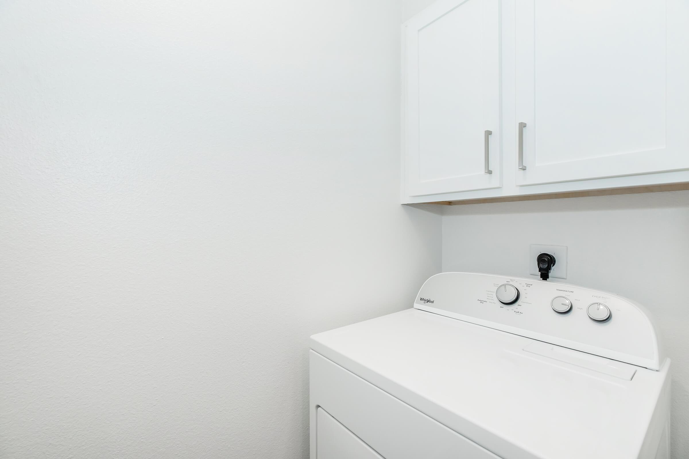 A white washing machine with control dials sits in a small laundry area. Above the machine, there are white cabinets against a plain wall. The space is well-lit and minimalistic, creating a tidy appearance.