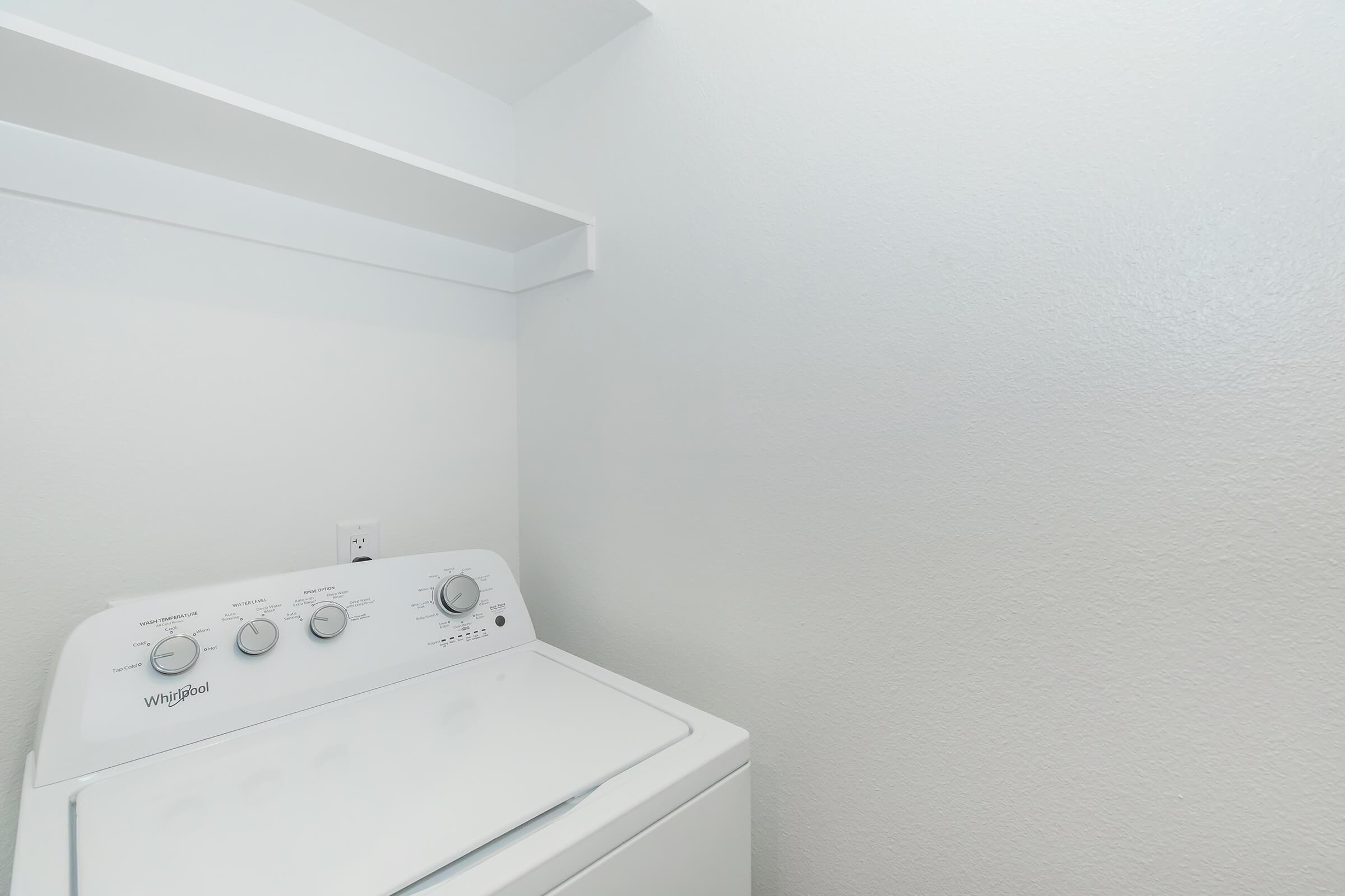 A clean, bright laundry area featuring a white Whirlpool washing machine with control dials. There is a wall-mounted shelf above the machine, and the walls are painted a light color, creating a spacious and uncluttered environment.