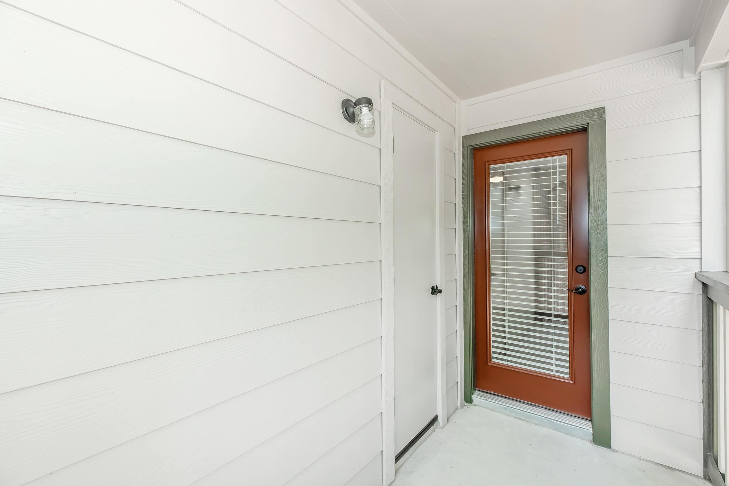 A well-lit entryway featuring white wooden paneling, a glass door with vertical blinds, and a dark-colored door adjacent to it. The area has a clean, modern aesthetic, with natural light illuminating the space.