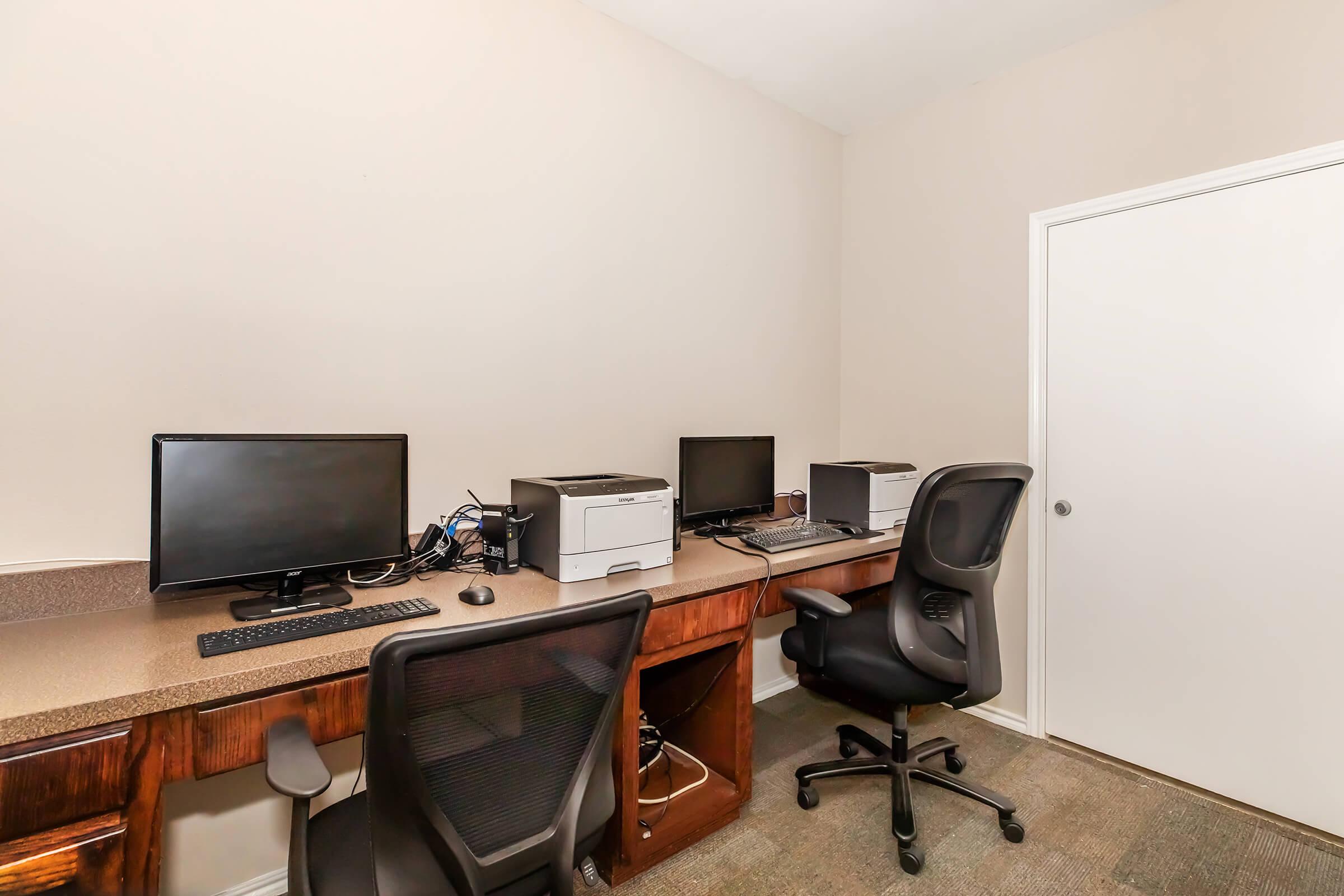 Two desks with computers and printers in a minimalist workspace. Each desk has a black office chair, and the walls are a neutral beige color. A closed white door is visible in the background. The overall atmosphere is clean and organized, suitable for a quiet working environment.