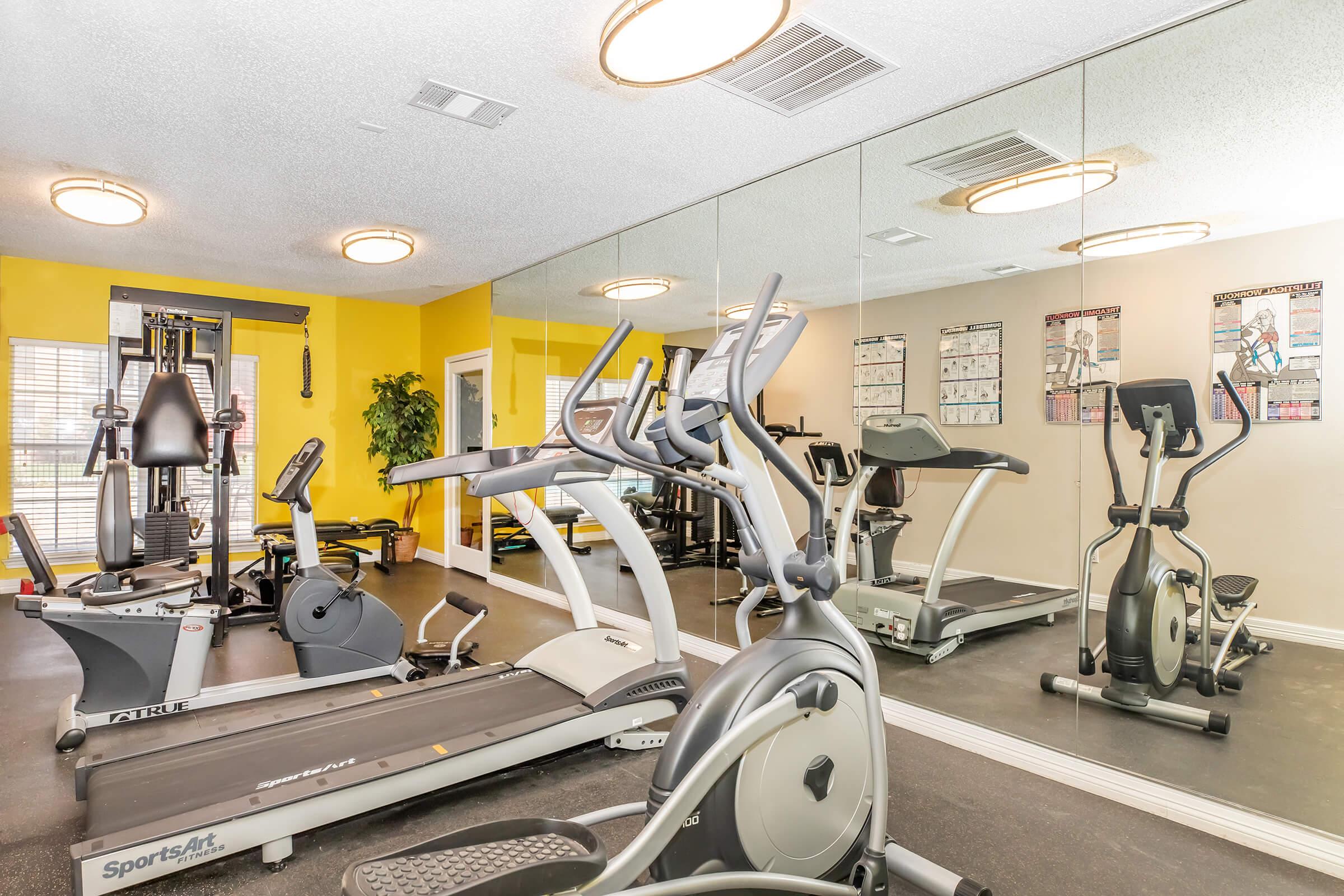 A well-equipped gym with exercise machines, including a treadmill and elliptical, set against a vibrant yellow wall. Mirrors reflect the workout equipment, creating an open and inviting space. Two framed posters with workout exercises are visible, enhancing the fitness environment.