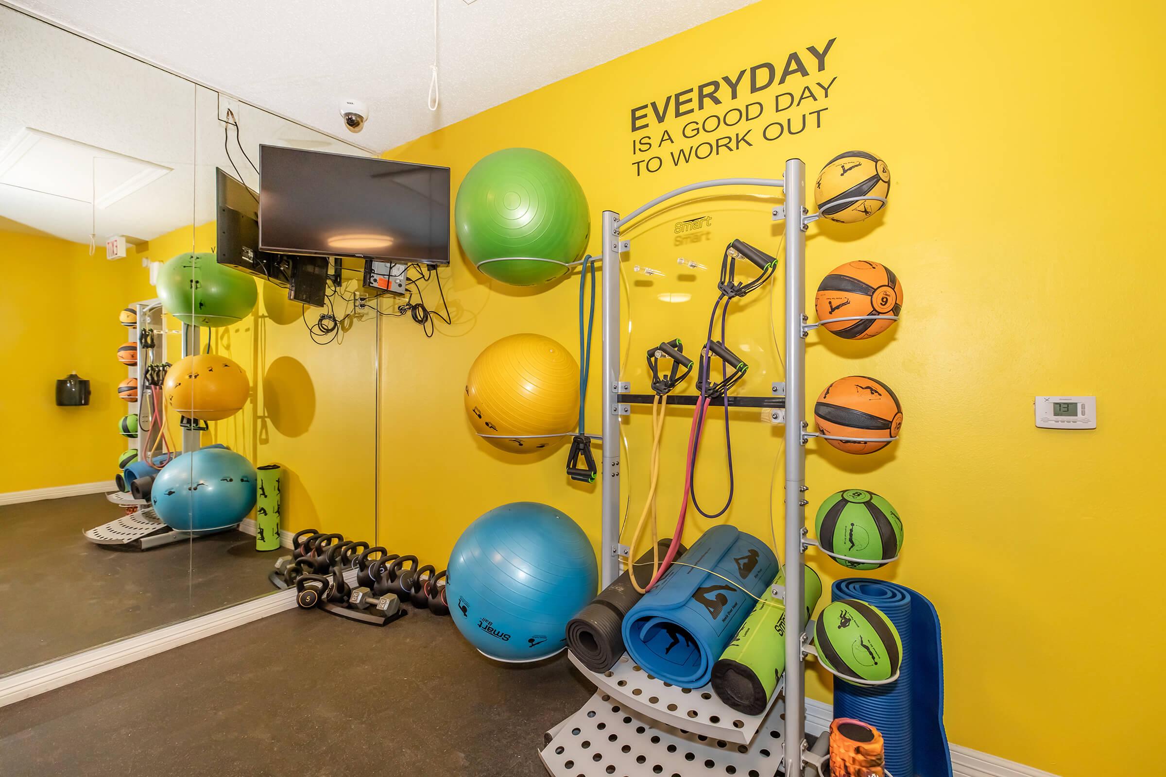 A brightly colored fitness room featuring exercise equipment including stability balls, resistance bands, and yoga mats. The wall is painted yellow with the motivational phrase "EVERYDAY IS A GOOD DAY TO WORK OUT." A mirror reflects the equipment, and there’s a television mounted on the wall.