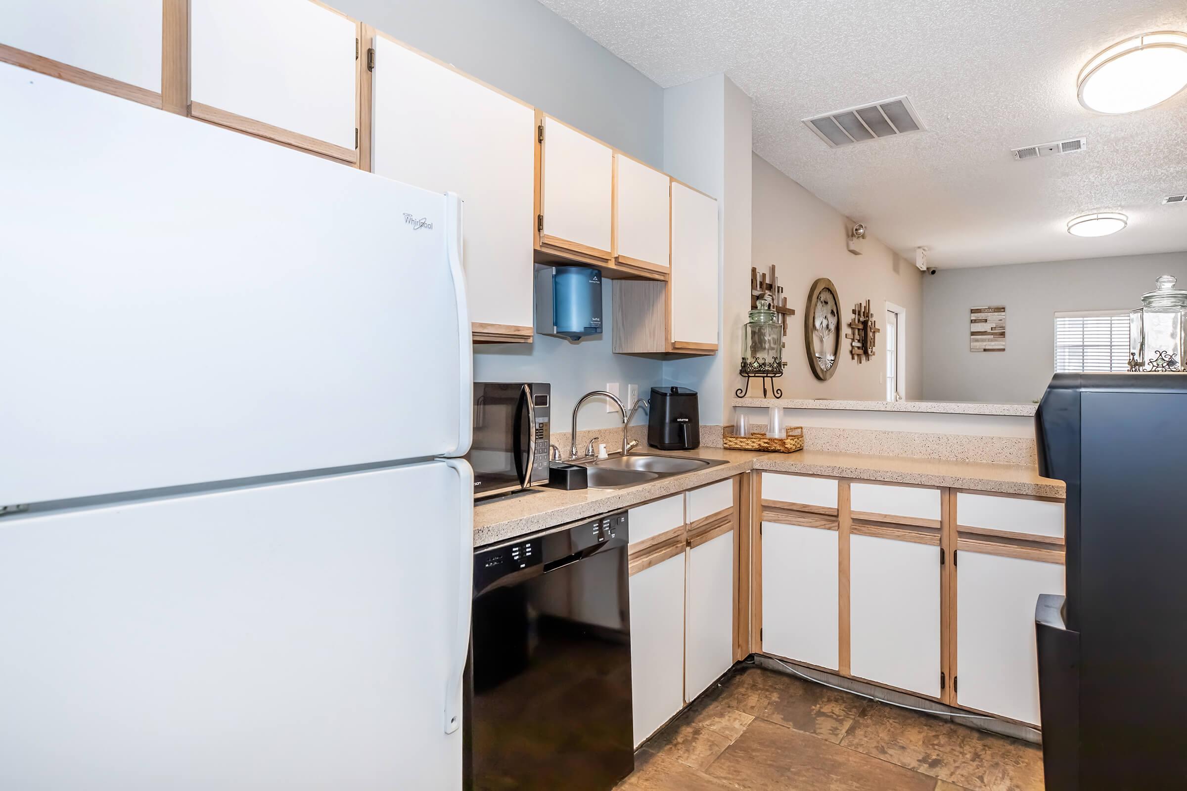 Modern kitchen featuring a white refrigerator, black dishwasher, and sink with stainless steel faucet. Light-colored cabinetry with wooden accents complements the beige countertop. The space is well-lit with overhead lighting and has a neutral color palette, creating an inviting atmosphere.