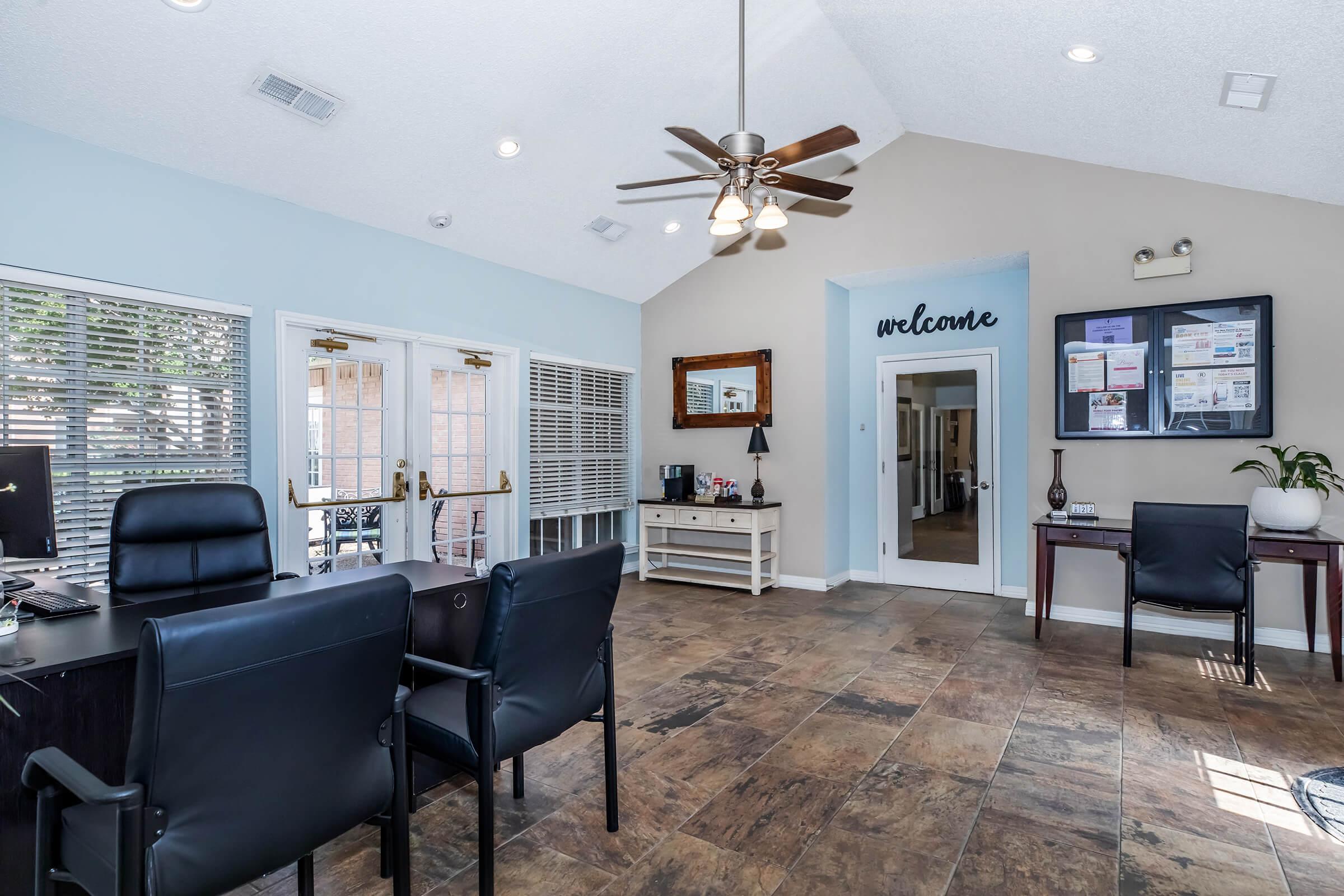 Interior view of an office reception area with two black chairs in the foreground, a wooden table with a plant, and a welcome sign on the wall. Large windows allow natural light in, and informational boards are displayed. The flooring is tiled, and the ceiling features a ceiling fan.