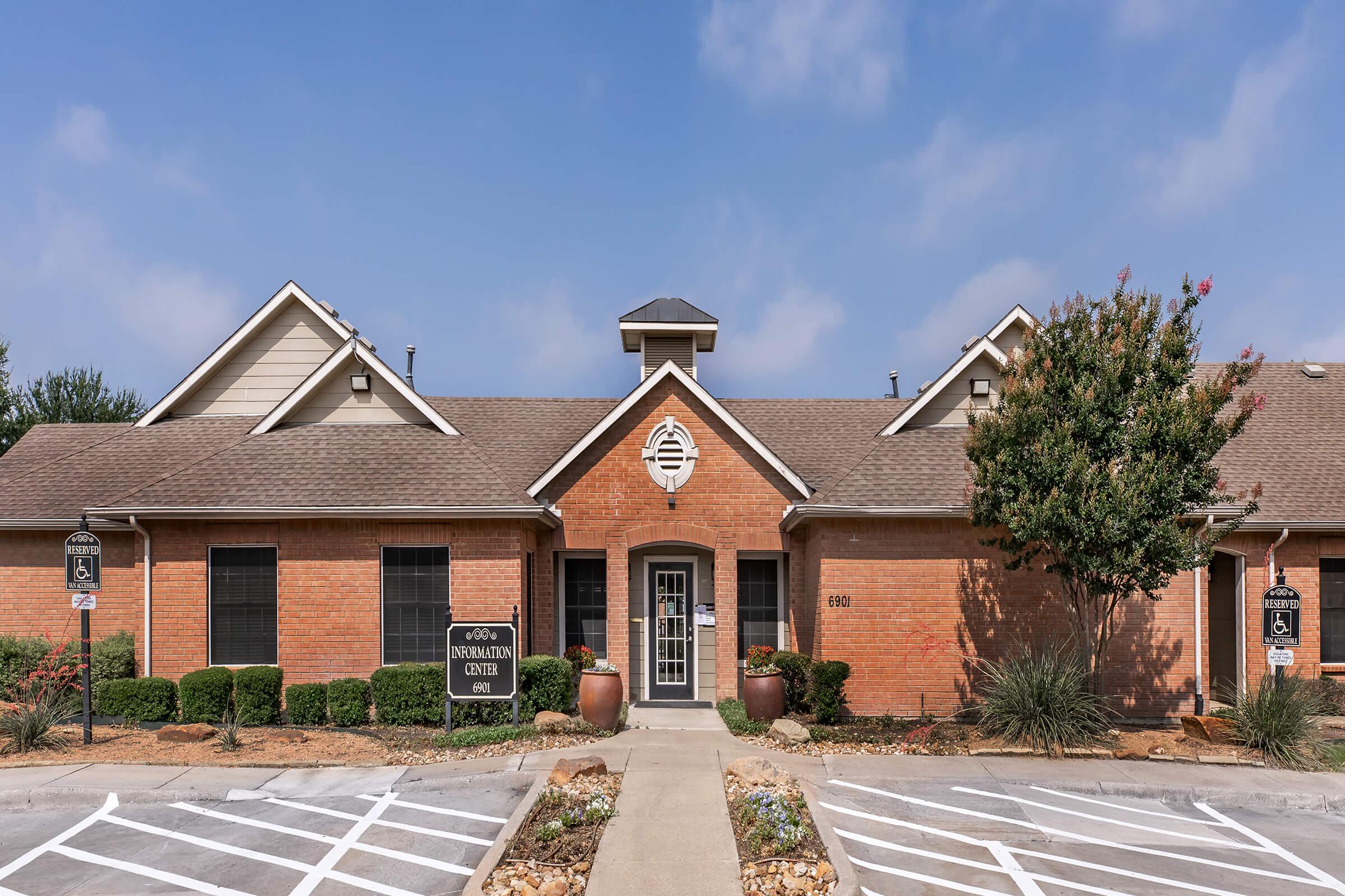 A single-story brick building with a sloped roof and a central entrance. The facade features decorative elements and the sign "Information Center" on the front. There are planters with shrubs and small trees, and a parking lot with marked spaces in front under a partly cloudy sky.