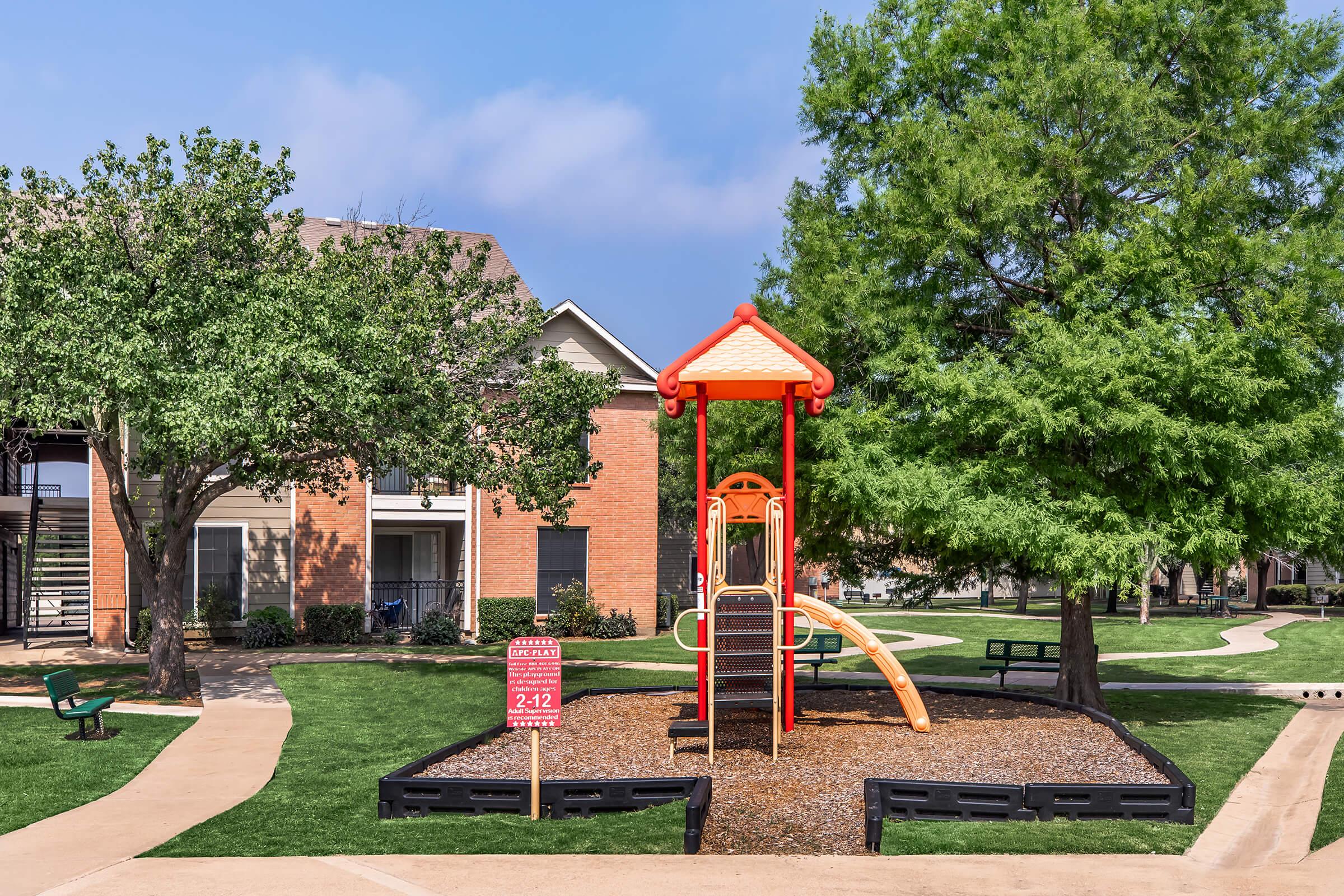 Colorful playground structure with a slide, surrounded by gravel, located in a grassy area with trees. Nearby are benches and pathways leading to apartment buildings. A sign is posted near the playground, indicating regulations or guidelines. Clear blue sky overhead.