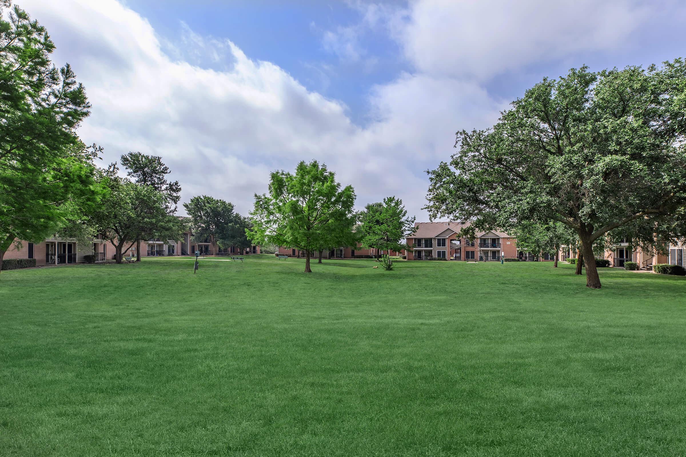 A wide, grassy area surrounded by trees and residential buildings under a partly cloudy sky. The landscape is lush and green, creating a serene and inviting outdoor environment.