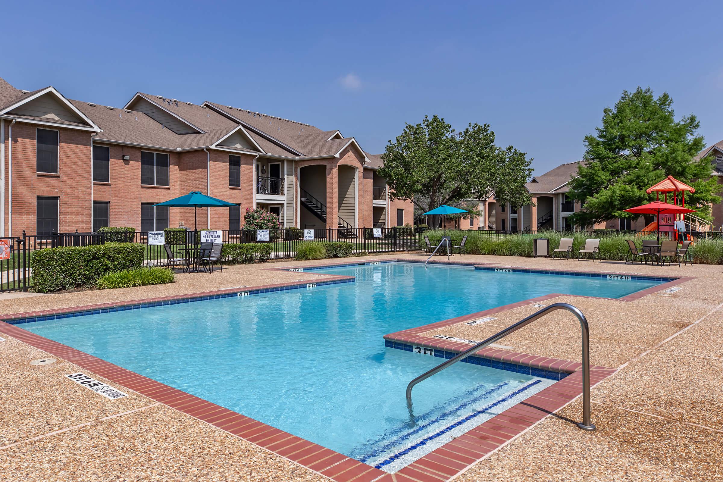 A bright outdoor swimming pool surrounded by a patio area, lounge chairs, and shaded seating under colorful umbrellas. Red brick apartment buildings in the background, along with well-maintained landscaping featuring trees and shrubs, create a relaxing community atmosphere on a sunny day.