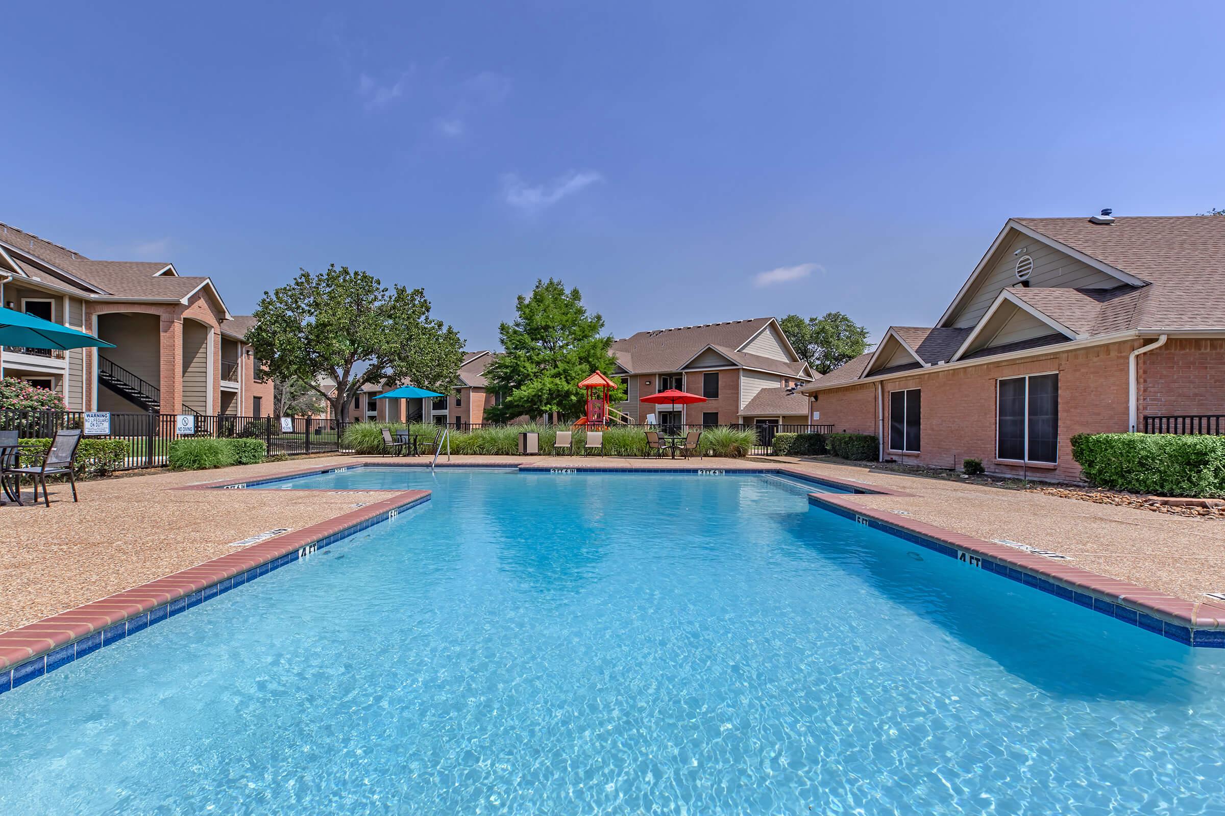 A clear blue swimming pool with a light brown concrete deck, surrounded by well-maintained grass and shrubs. In the background, there are several apartment buildings with balconies. Colorful umbrellas in red and teal are set up around the pool area, under a bright blue sky.
