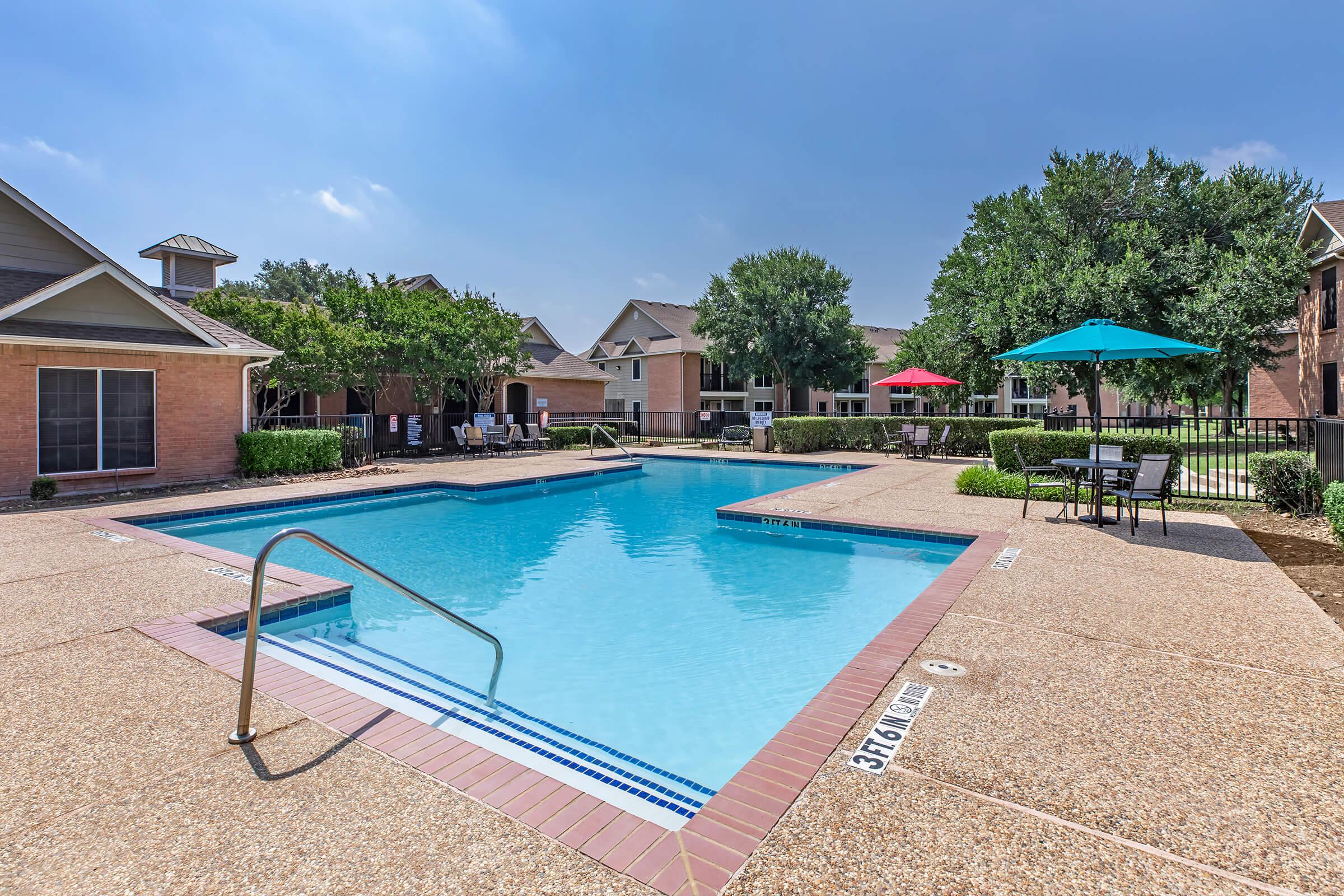 A clean, inviting swimming pool surrounded by a patio area. There are lounge chairs and tables with umbrellas, alongside green trees and nearby buildings. The sky is clear with a few clouds, creating a bright atmosphere.