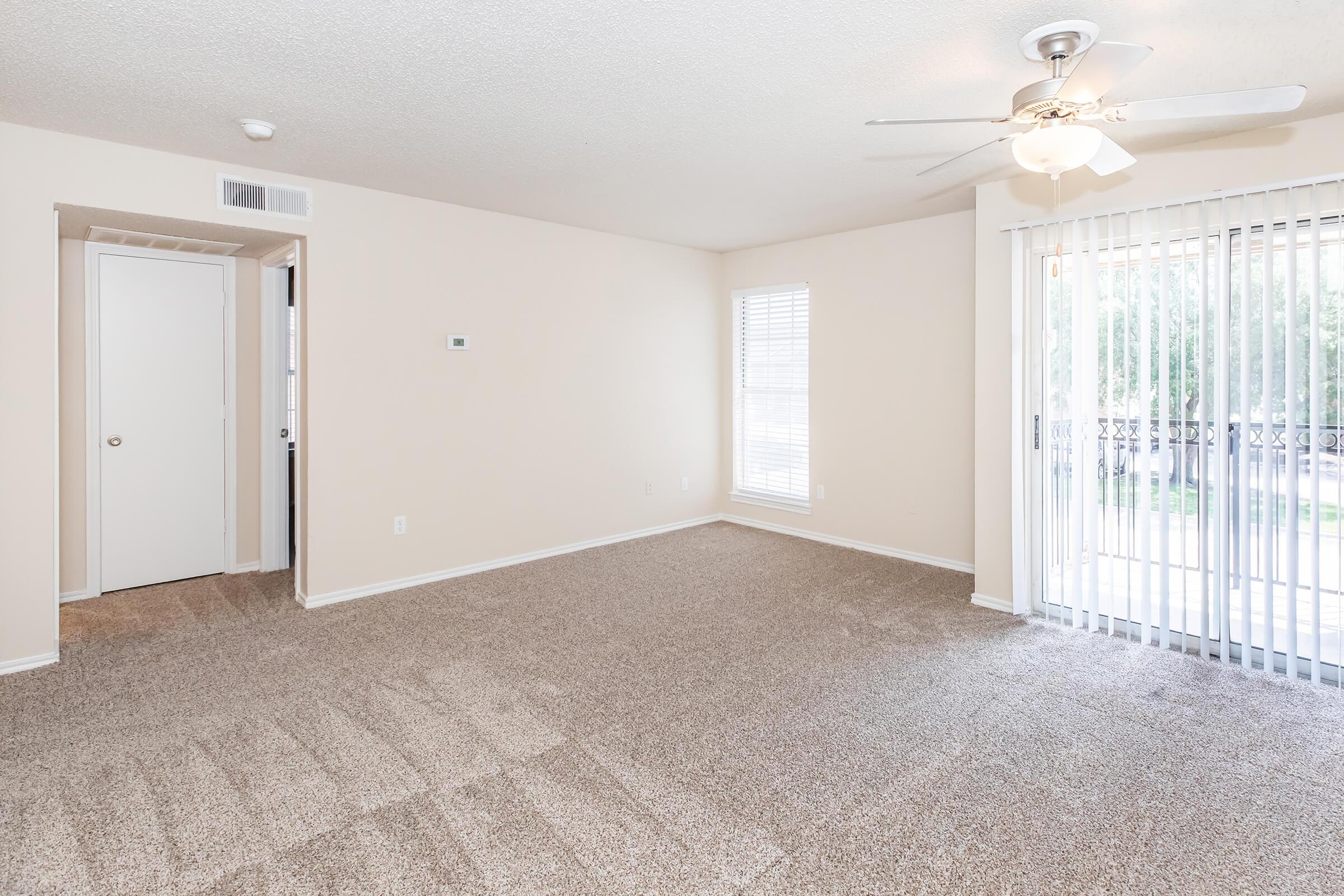 A spacious, empty living room featuring light beige walls and plush carpeting. There's a ceiling fan for airflow, and a set of sliding glass doors leading to an outdoor balcony. One wall includes a small window with vertical blinds, and a closet door is visible on the left side of the room.