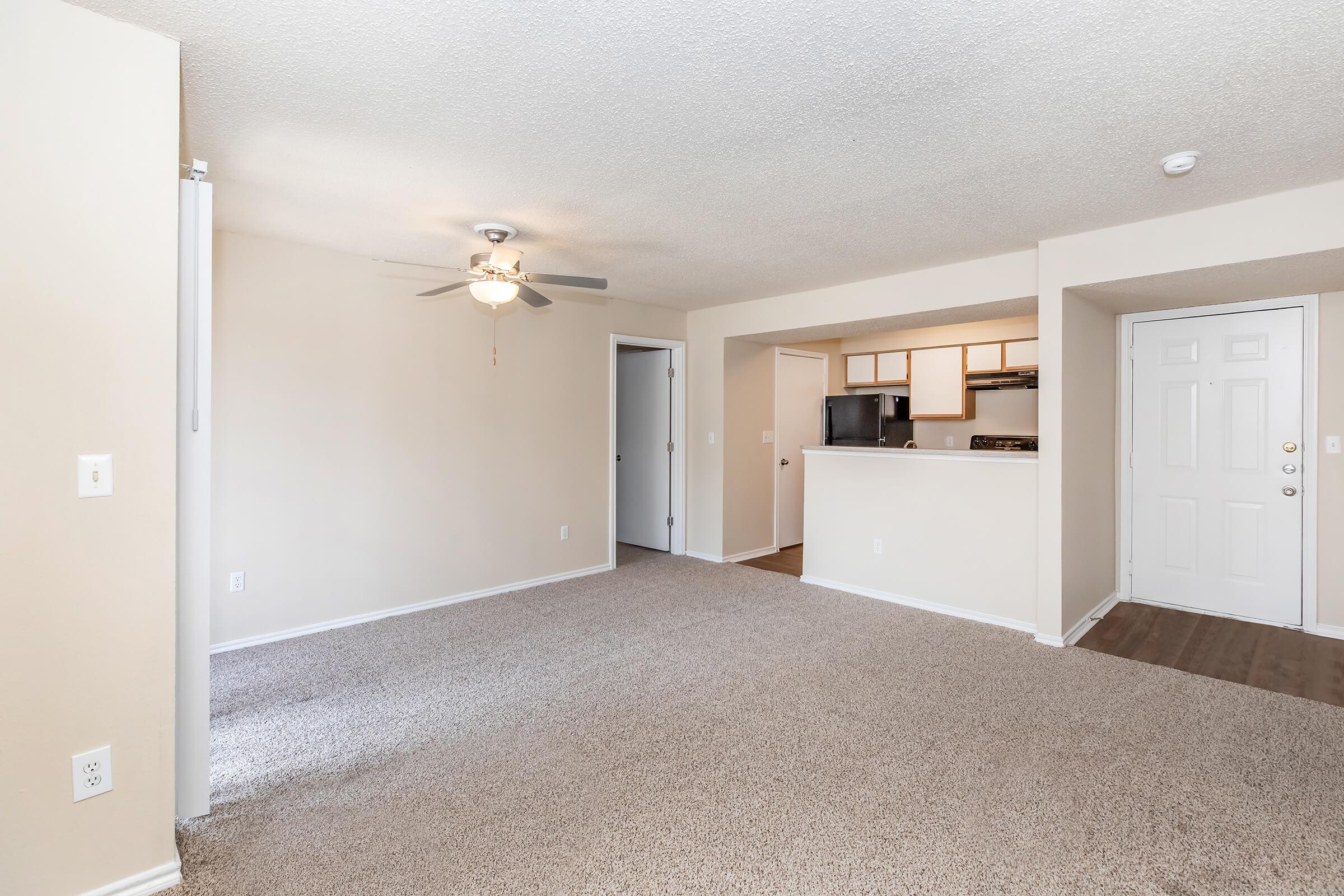 A spacious, well-lit living area featuring beige carpeting, a ceiling fan, and light-colored walls. To the right, a doorway leads to a kitchen with dark cabinetry and appliances. An entrance door is visible on the far right, while another door leads to a hallway.