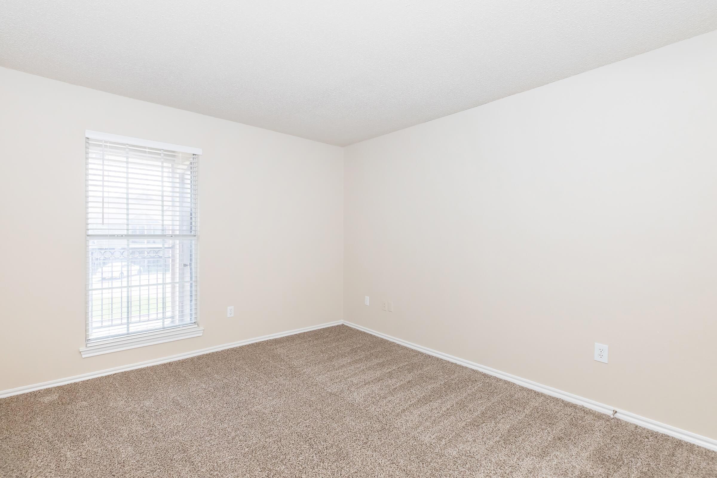 A vacant room with light beige walls and plush beige carpet. The window has white blinds and allows natural light to enter, illuminating the space. The room is empty, providing a blank canvas for decorating or furnishing.