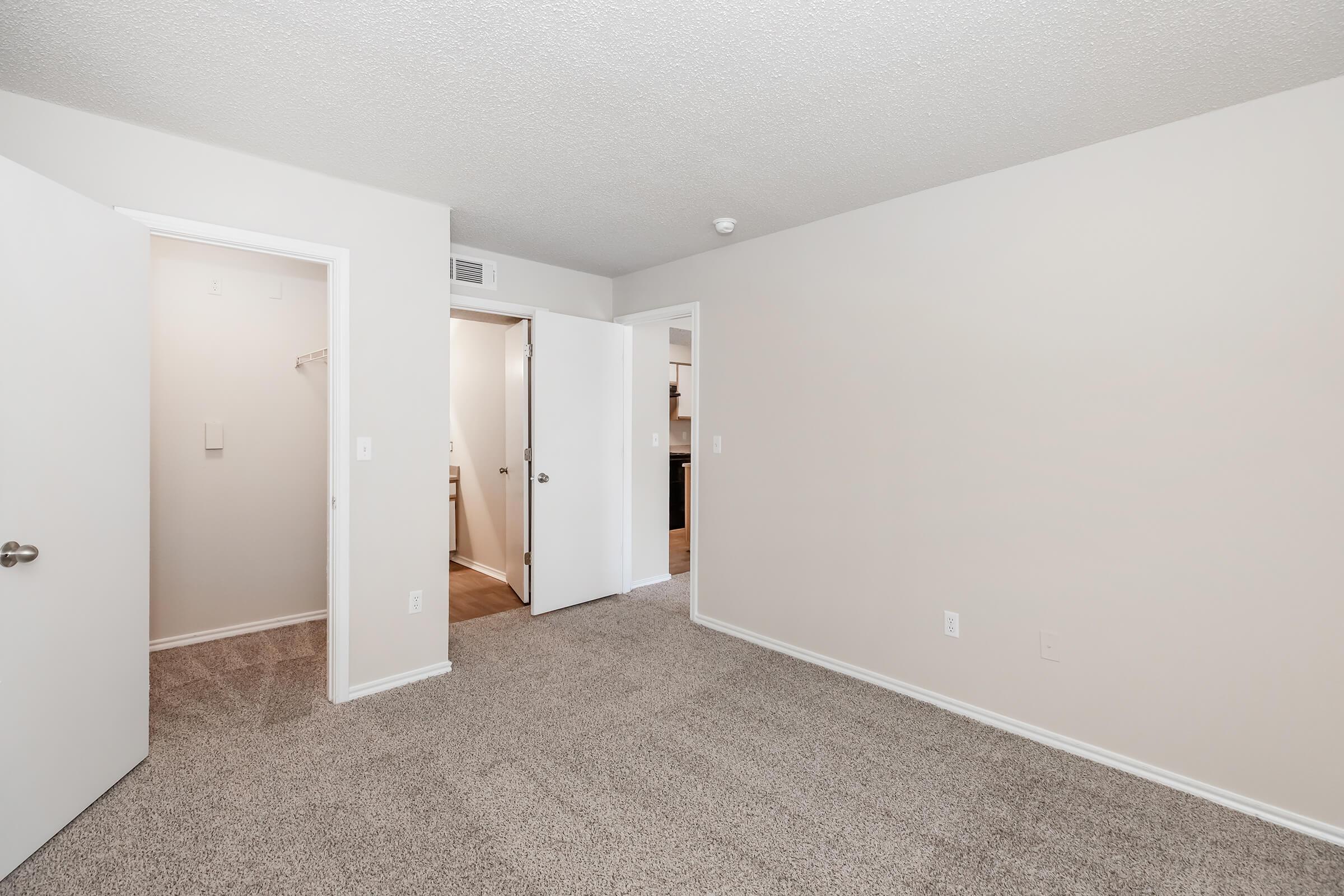 A spacious, empty room with light beige walls and plush carpet. Two doors on the left lead to closets or other areas, while a third door on the right opens to another room. The overall atmosphere is bright and inviting, ideal for customization or decoration.