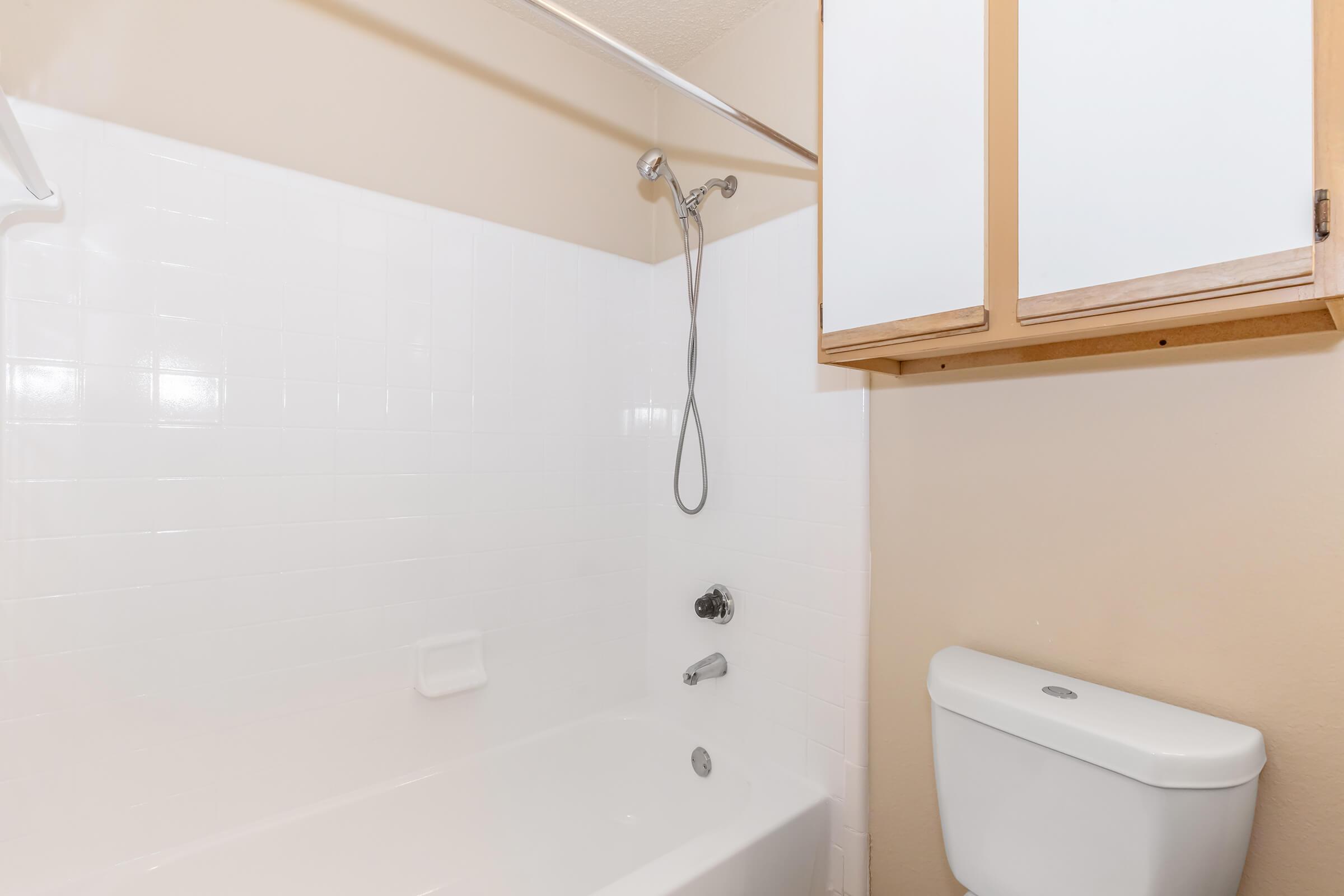 A clean bathroom featuring a white bathtub with a showerhead, a wall-mounted shower caddy, and a toilet next to a wooden cabinet above. The walls are light-colored, and the space is well-lit, creating a simple and functional atmosphere.