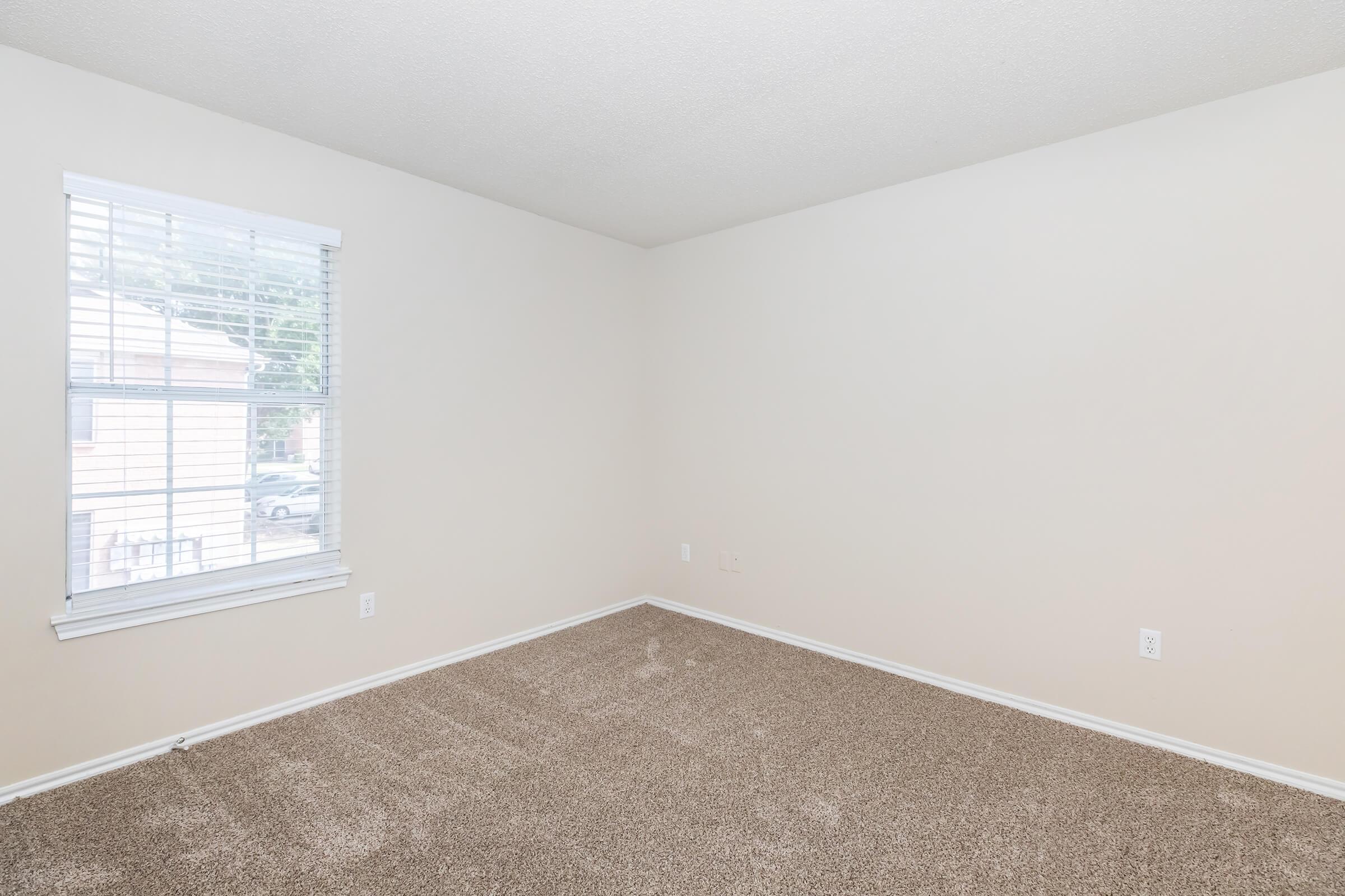 A vacant room with light beige walls and a window covered with white blinds. The floor is covered in light brown carpet, and there are no furniture or decorations. The setting appears bright and airy with natural light coming in through the window.