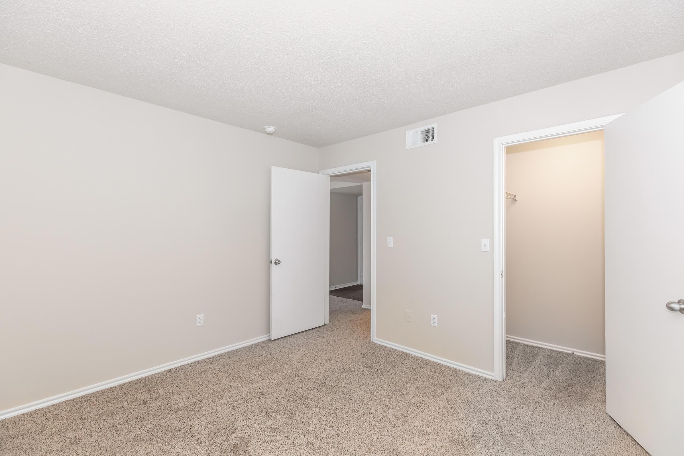 A spacious, neutral-colored bedroom with carpeted flooring, featuring two open doors leading to adjacent rooms. The walls are painted light, and there is a ceiling with a textured finish. One door leads to a closet area. Natural light may be present, enhancing the room's simple and clean aesthetic.