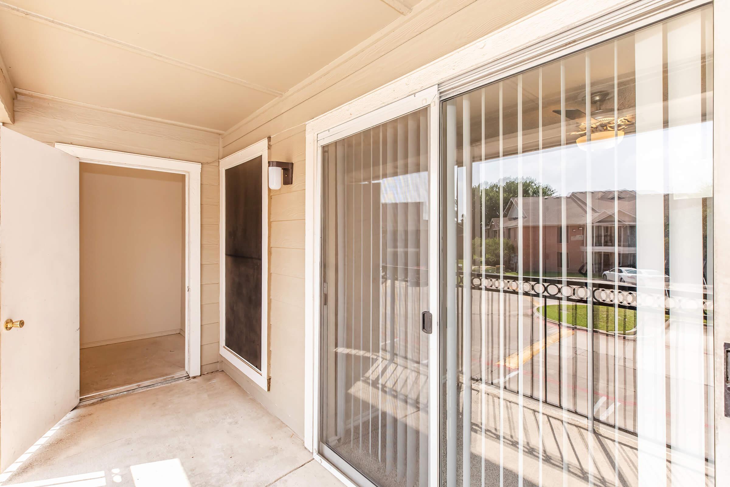 View of a porch area with a door on the left leading inside and sliding glass doors on the right. The porch has beige walls and a light fixture, with a glimpse of greenery and parked cars visible through the glass doors. The space is empty and well-lit.