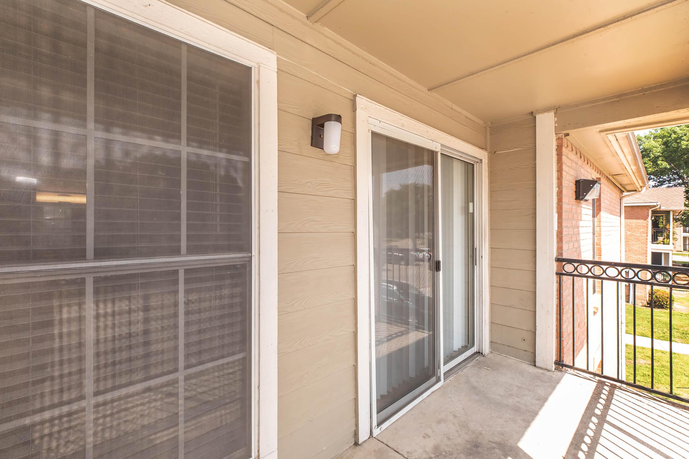 A balcony view featuring sliding glass doors and a light fixture on the wall. The exterior is painted in light colors with a railing and a view of green grass and trees in the background. The adjacent wall has a window with blinds.