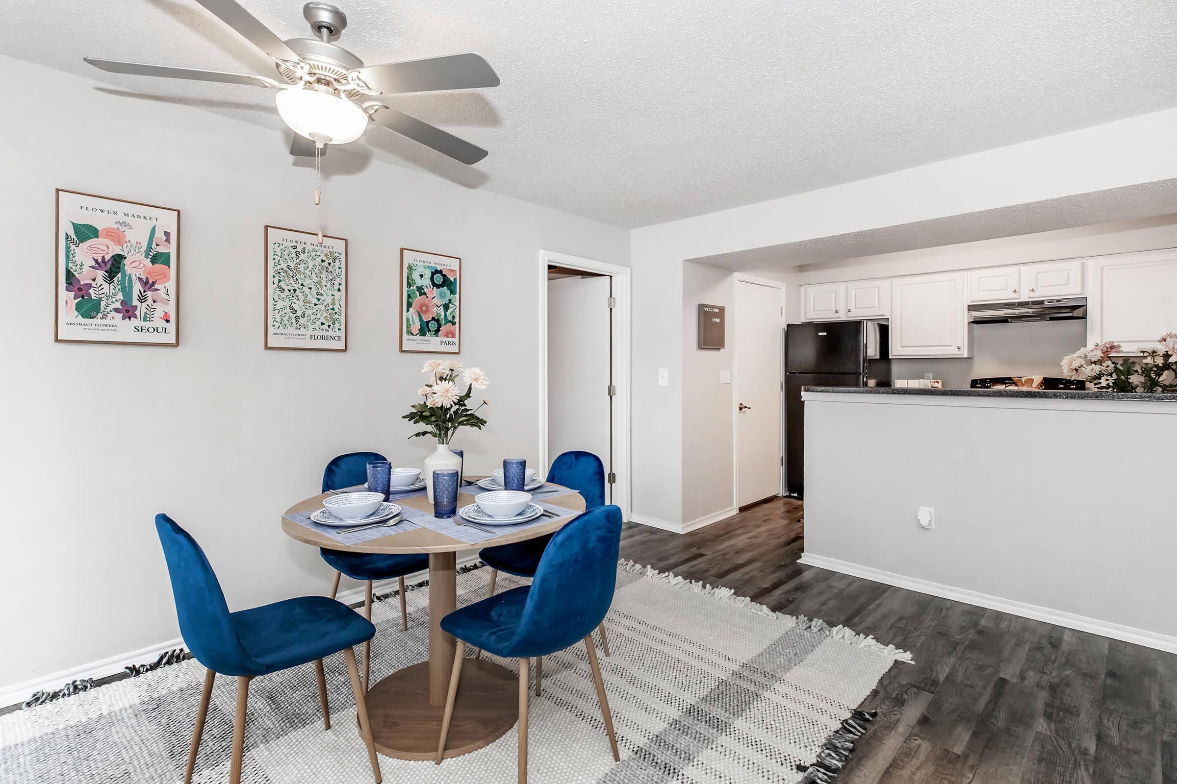 A modern dining area featuring a round wooden table set with blue plates and glasses, surrounded by four blue upholstered chairs. A ceiling fan is visible above, and two floral wall art pieces hang on the light-colored walls. The kitchen is partially visible in the background.
