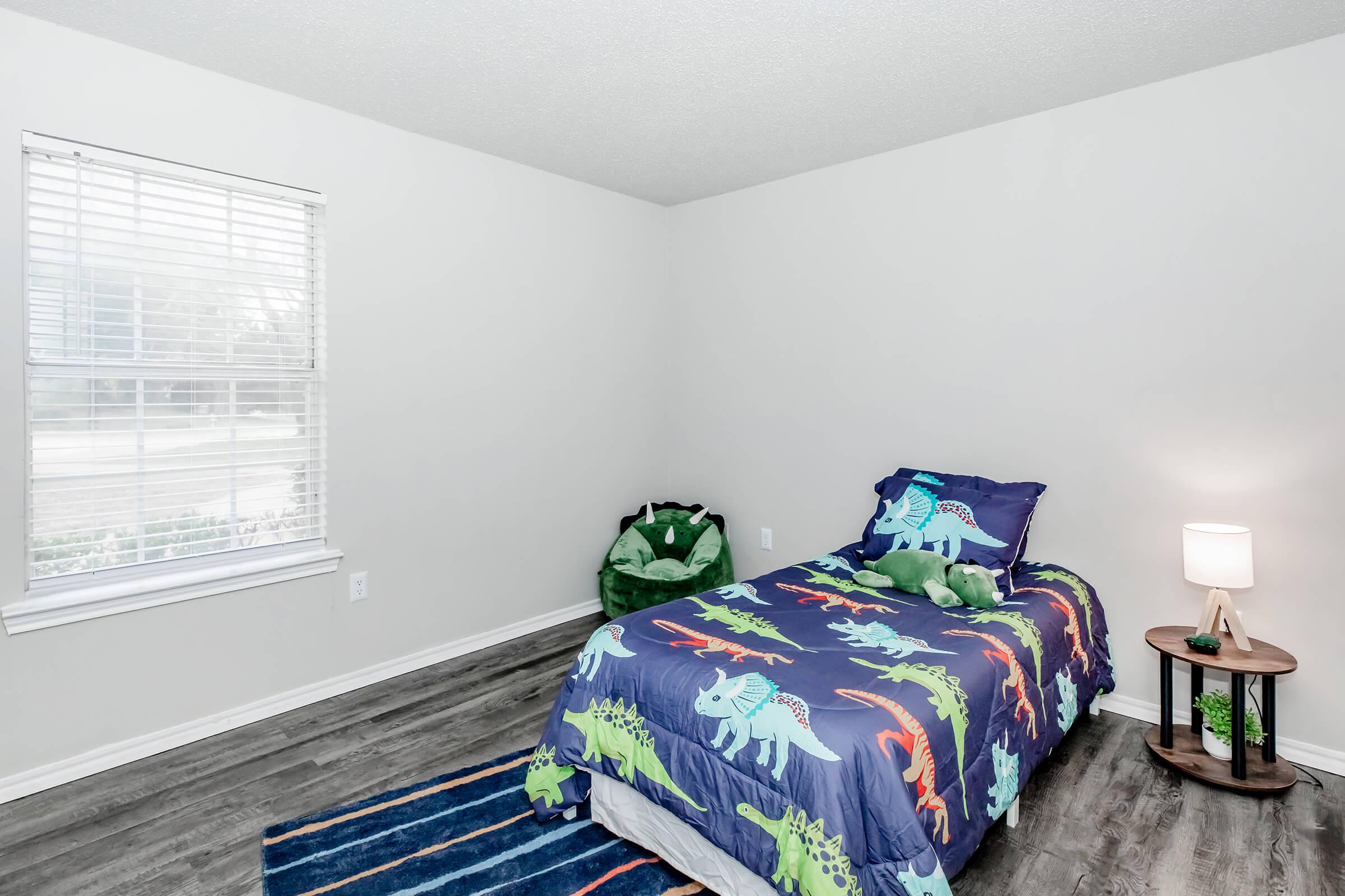 A simple bedroom featuring a single bed with dinosaur-themed bedding, a green plush dinosaur toy, a small bedside table with a lamp, and a dark blue area rug. Natural light enters through a window with white blinds, and the walls are painted a soft gray. A green bean bag chair is positioned in the corner.