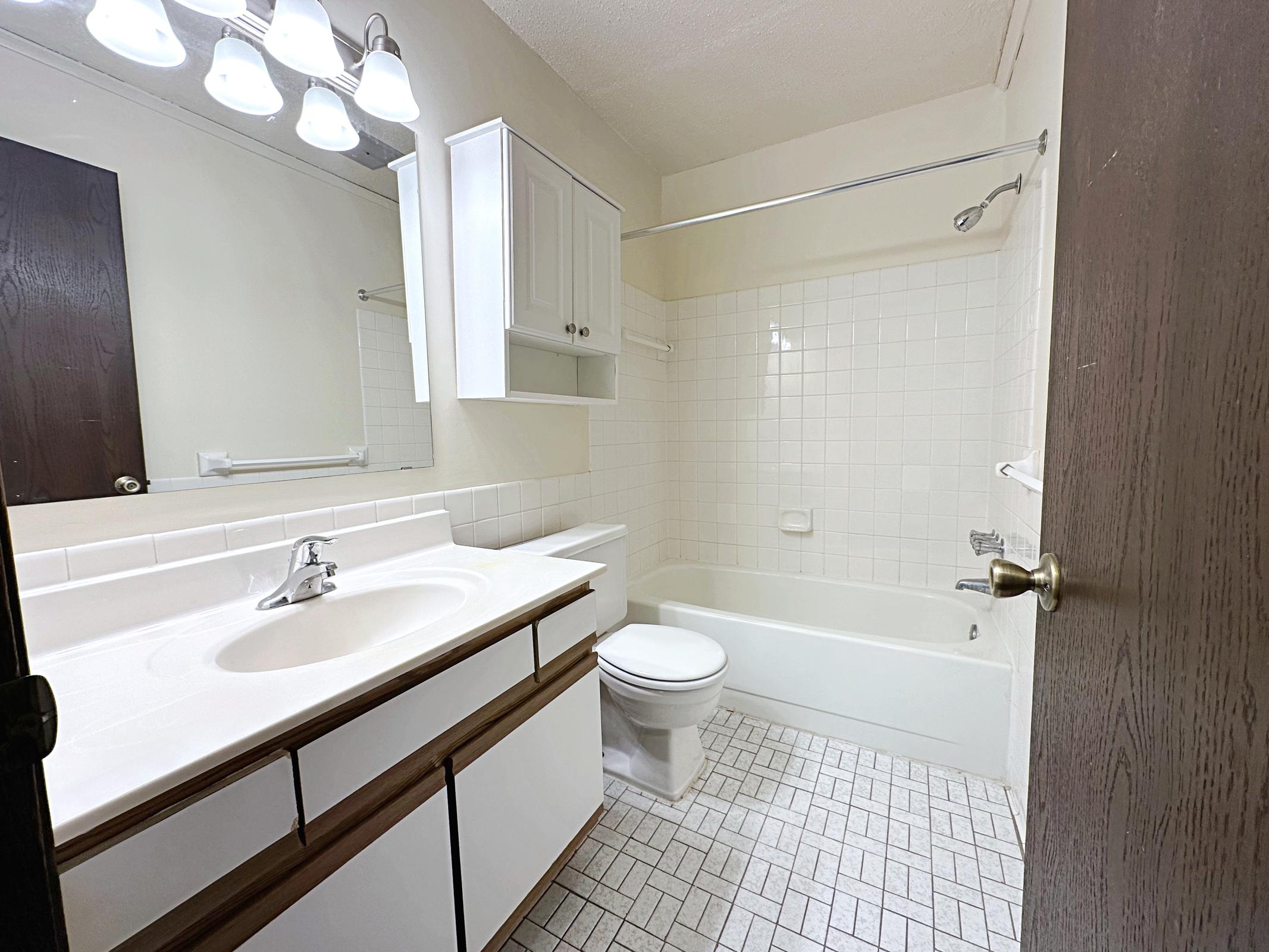 A clean, well-lit bathroom featuring a white tiled tub, a toilet, and a sink with a light-colored countertop. Above the sink is a mirror and cabinet. The floor has a textured tile design, and there are four light fixtures above the mirror. A wooden door is partially visible, leading to the hallway.