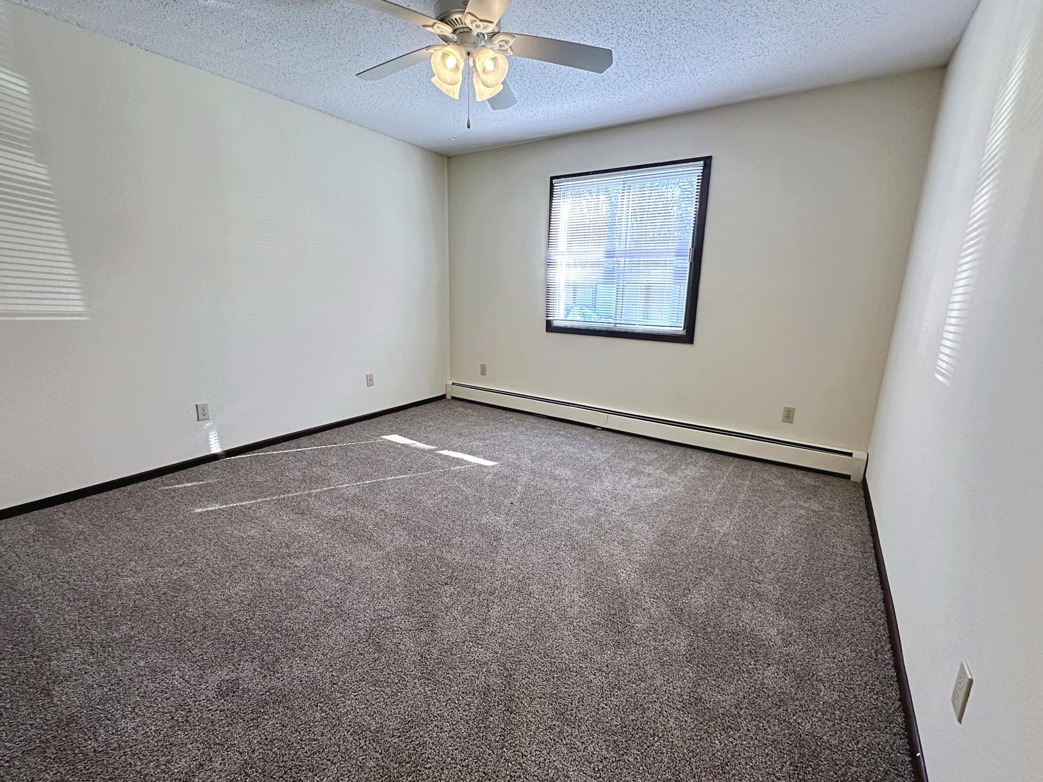 A vacant room with light beige walls and a dark carpet. It features a ceiling fan with four lights and a window that allows natural light to enter. There are no furnishings, making the space appear open and ready for decoration. The room's corners create an angled appearance due to the wall layout.