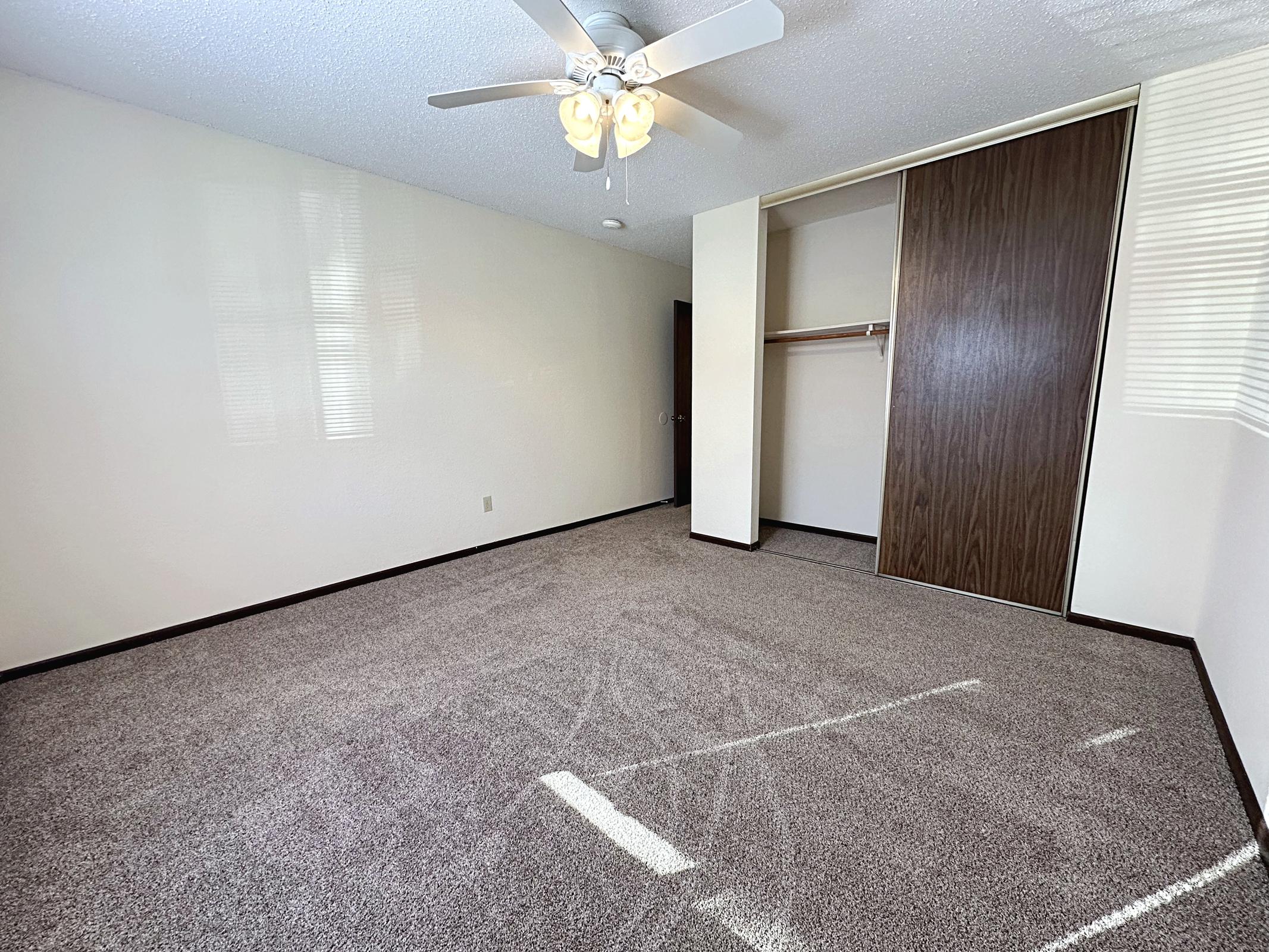 Empty room with light-colored walls and a ceiling fan. The floor is covered with carpet, and there is a closet with a sliding door on the right. Natural light comes through the window, creating a bright and spacious atmosphere.