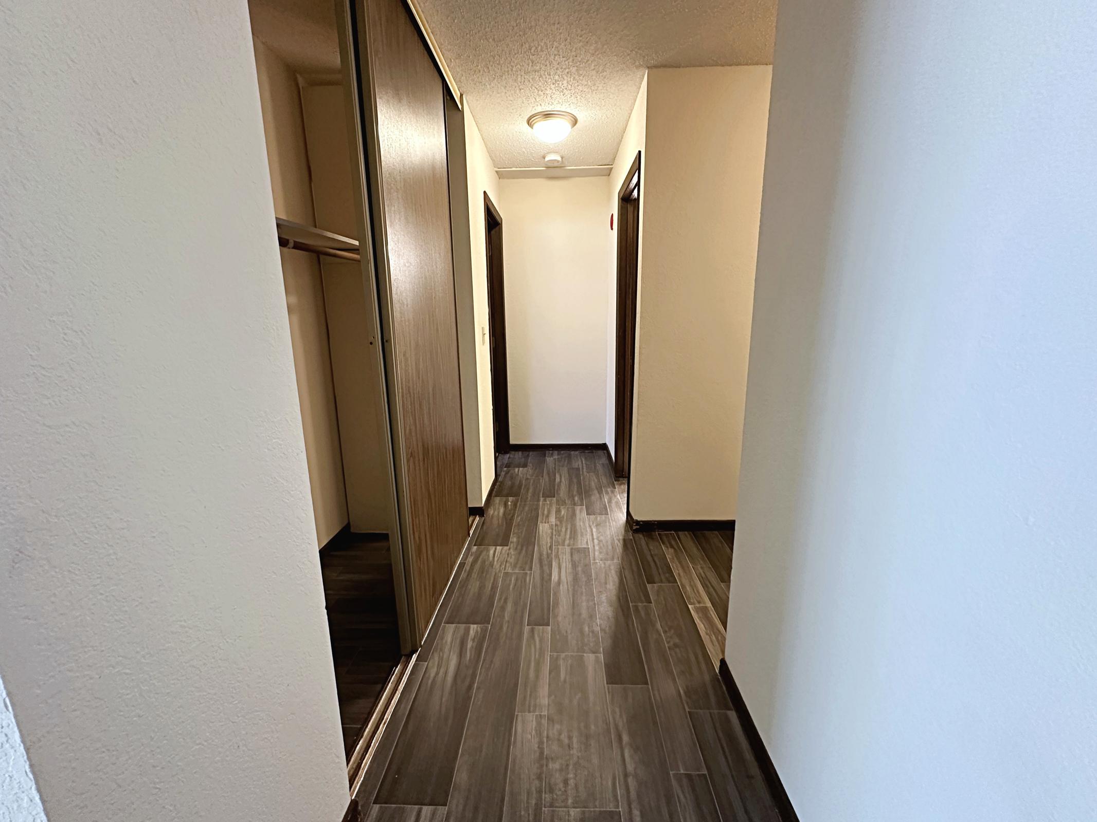 A well-lit hallway with dark tiled flooring and light-colored walls. On one side, there are built-in closets with wooden doors, and opposite, there are several doorways leading to other rooms. The ceiling features a circular light fixture, enhancing the bright ambiance.