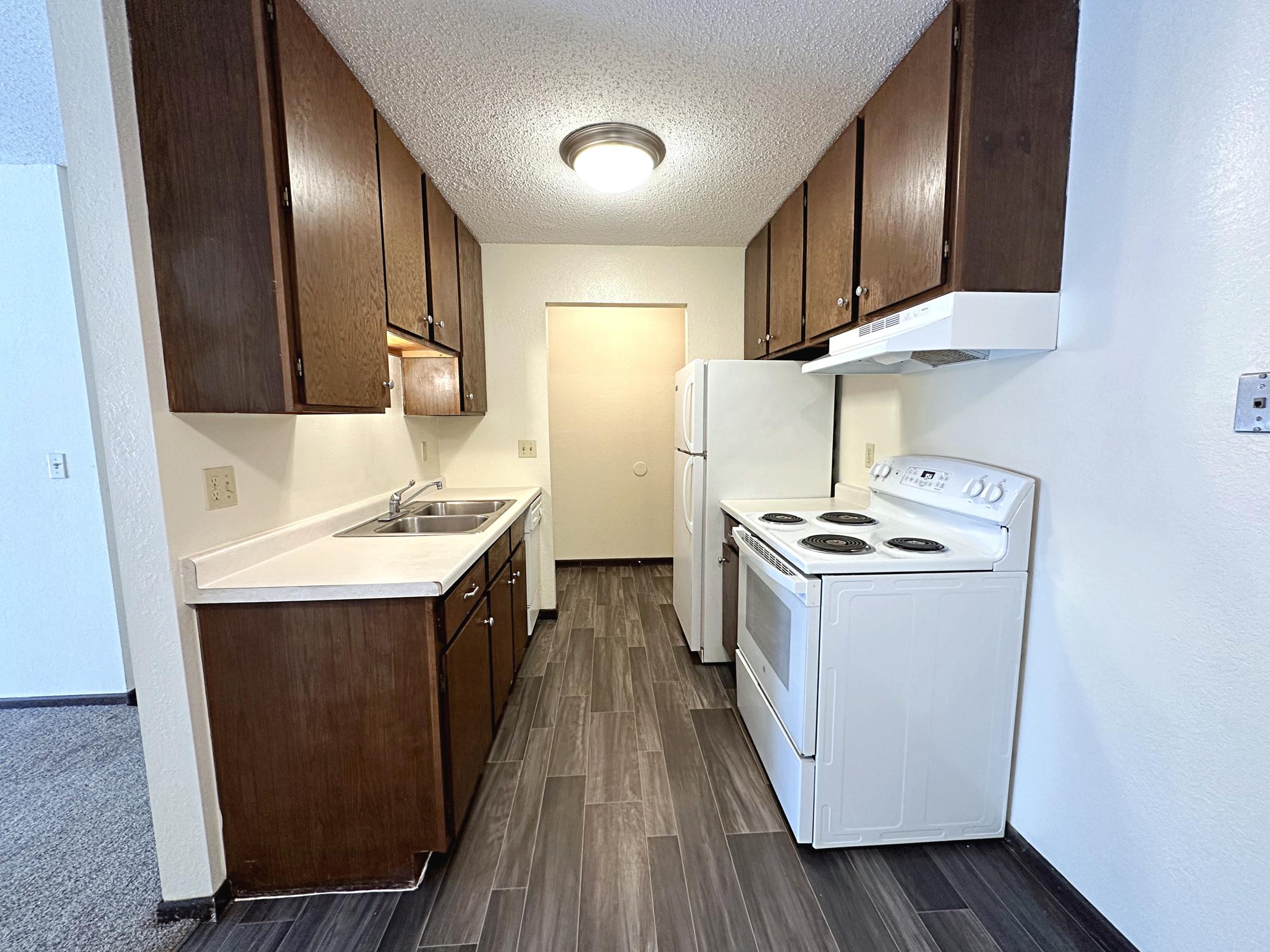 A clean, modern kitchen featuring dark wooden cabinets, white countertops, a double sink, and a white stove with oven. The space includes a refrigerator, an overhead light, and is designed with a contemporary tile floor. The adjacent doorway leads to another room, while natural light brightens the area.