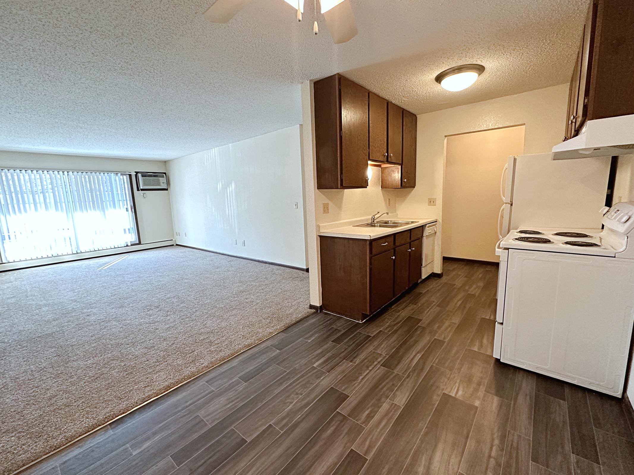 A spacious living area leads to a kitchen with wooden cabinets, a sink, and a stove. The flooring is dark tile, while the living room features carpet and a large window with vertical blinds. A wall-mounted air conditioner is visible, enhancing the comfort of the space.