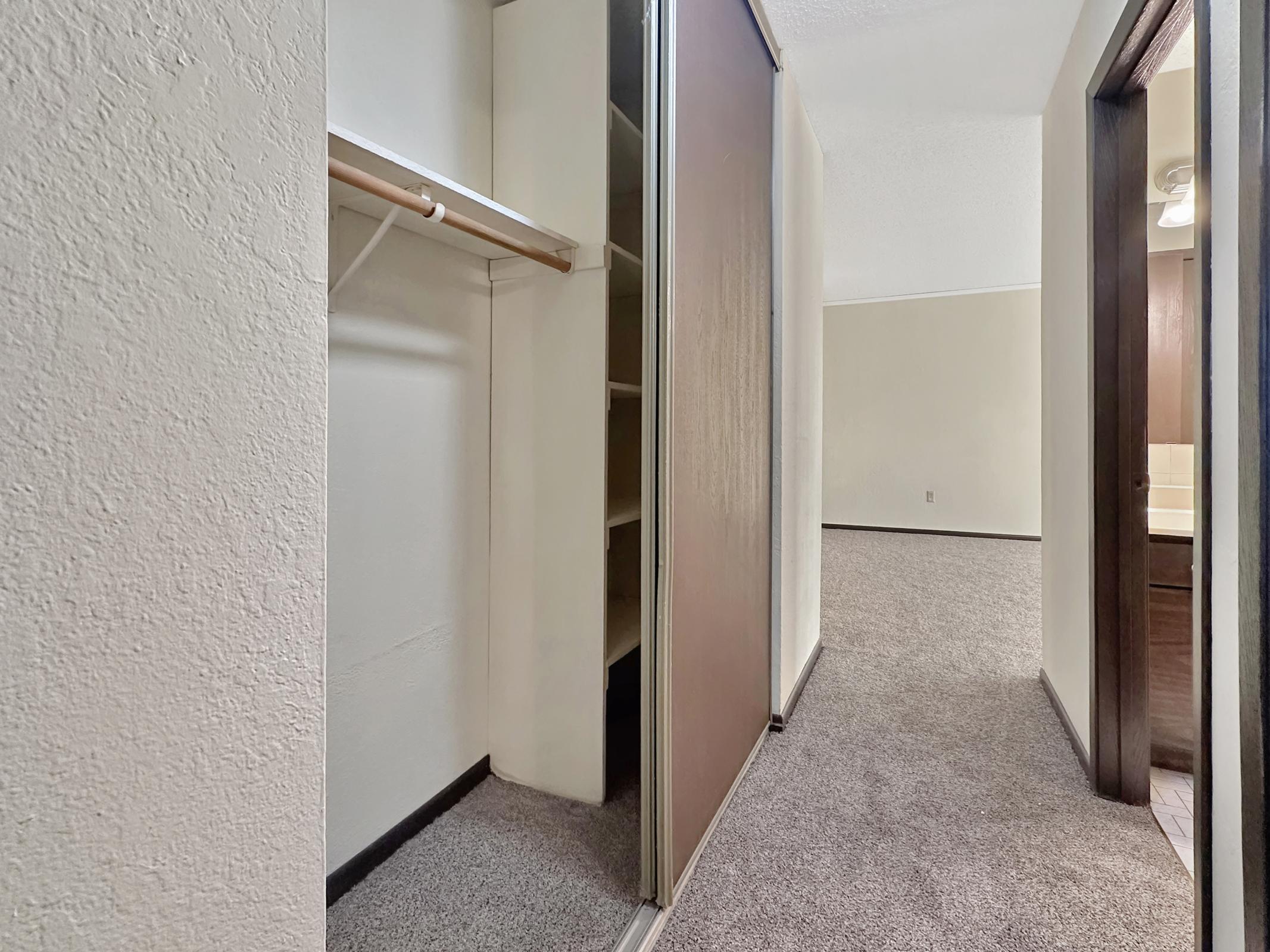 View of a small hallway featuring a closet with sliding doors on the left, partially open, revealing shelves inside. The right side shows an open doorway leading to another room. The flooring is carpeted, and the walls are painted in neutral tones, creating a bright and clean appearance.