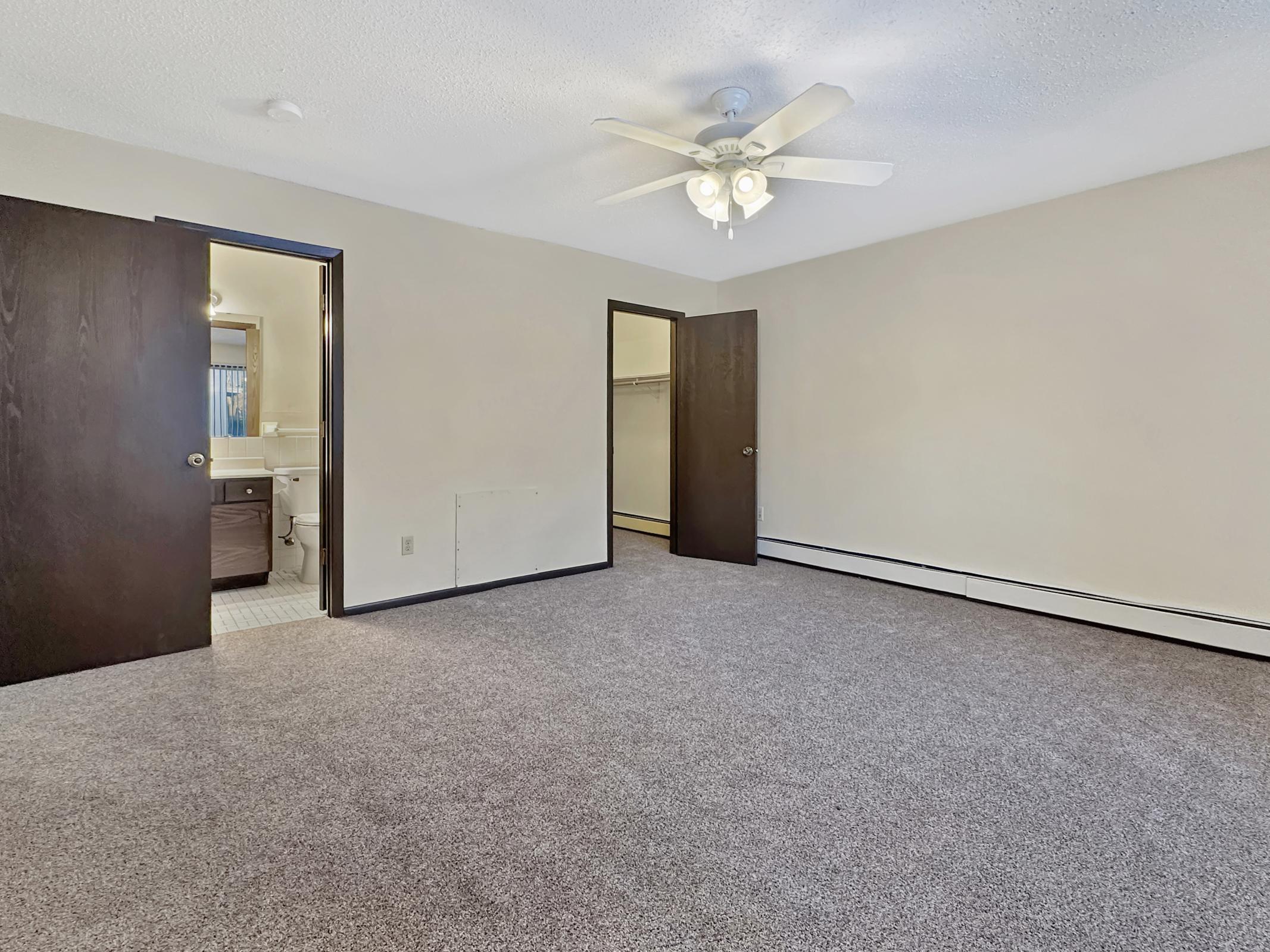 A spacious, empty room with light beige walls and gray carpet. There are two doors leading to other areas, one to a bathroom with visible fixtures. A ceiling fan hangs in the center, providing a bright, open feel to the space.