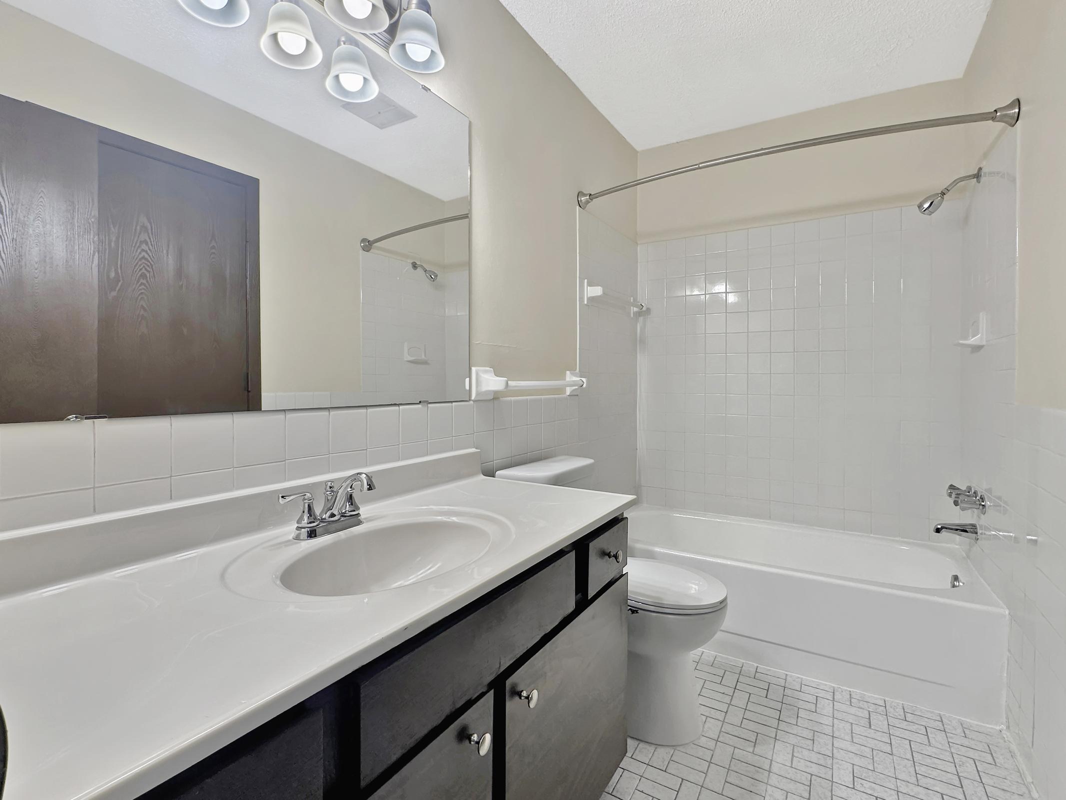 Interior view of a bathroom featuring a white bathtub and shower, a white sink with a mirror above, and dark cabinetry. The floor is tiled in a light pattern. There is a light fixture with multiple bulbs and a towel bar alongside the bathtub. The walls are painted a neutral color.
