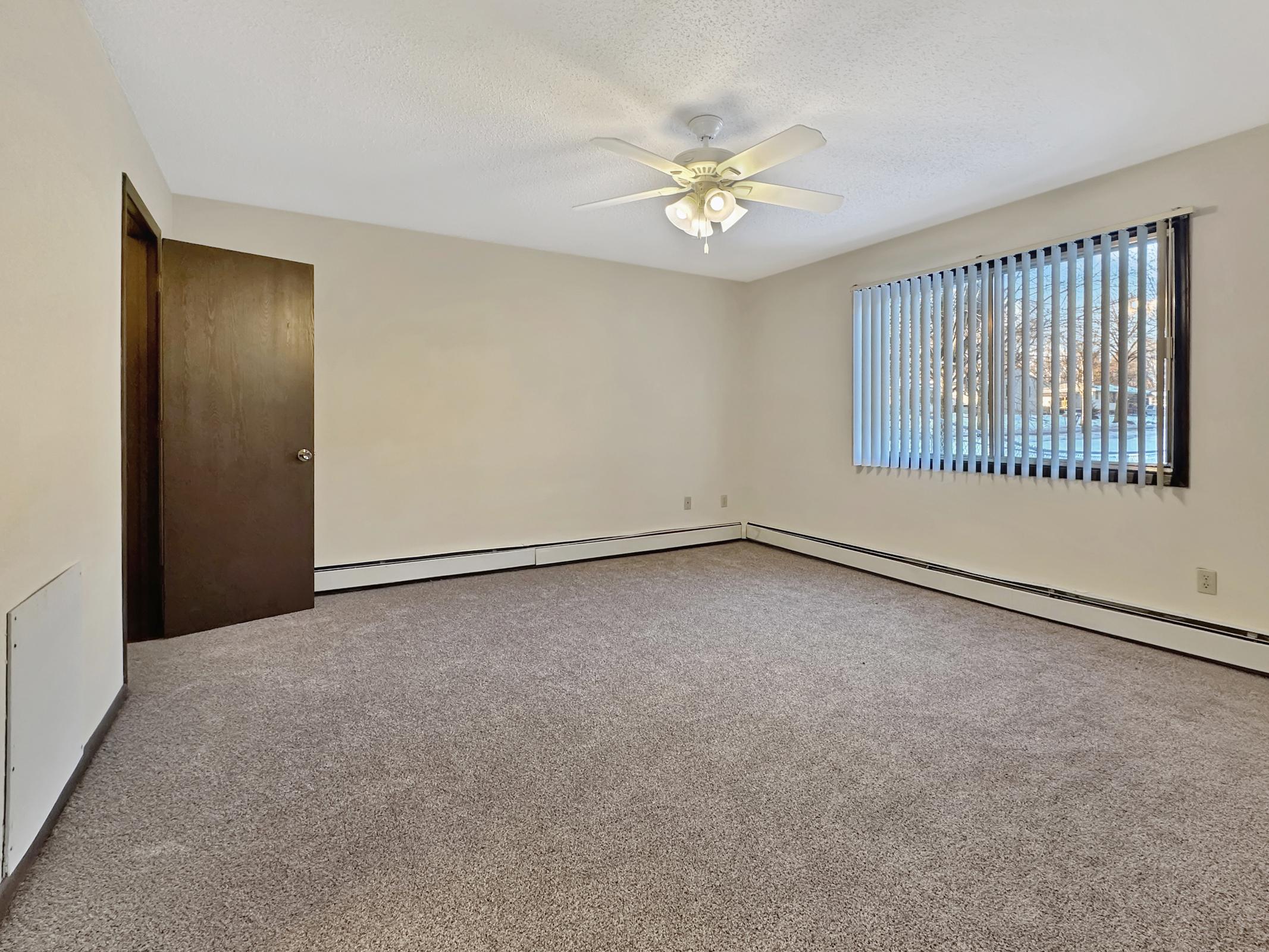A spacious, empty room featuring beige walls and light carpet. A ceiling fan is installed, and there is a large window with vertical blinds allowing natural light to enter. A closed brown door is visible. The room appears clean and ready for furnishing.