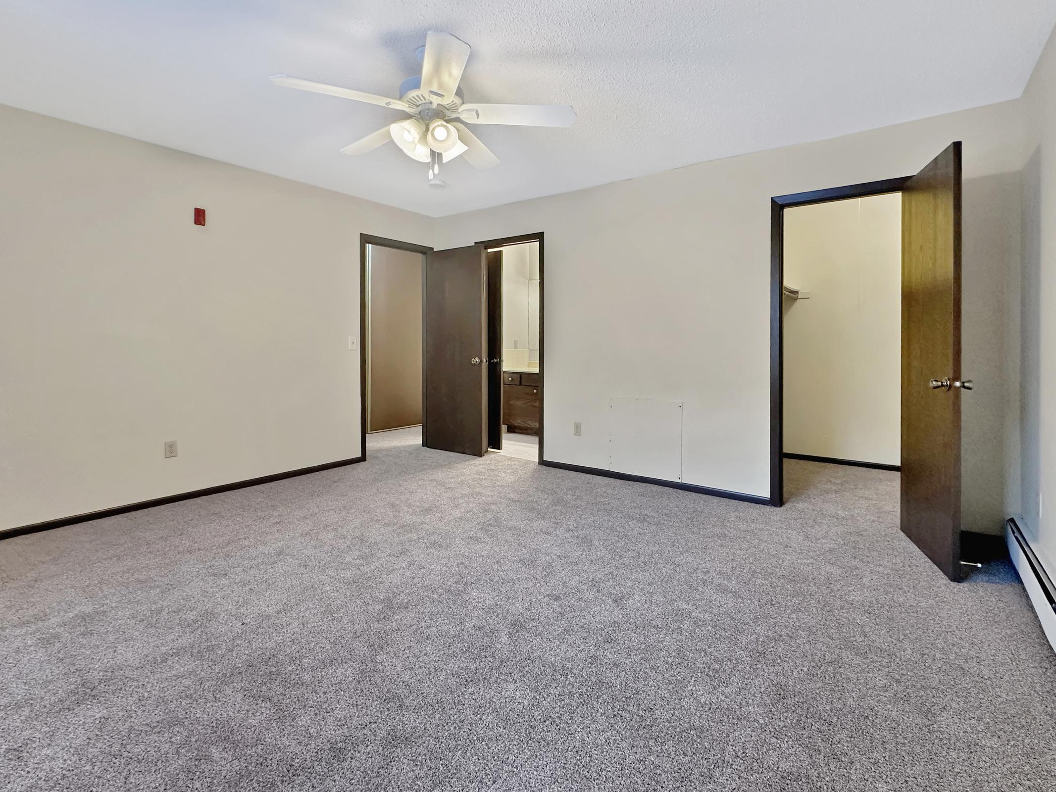 A spacious, empty room with light-colored walls and a ceiling fan. There are two doors leading to a bathroom and a closet. The floor is covered in carpet, and the space is well-lit with natural light. The overall atmosphere is clean and ready for decoration.