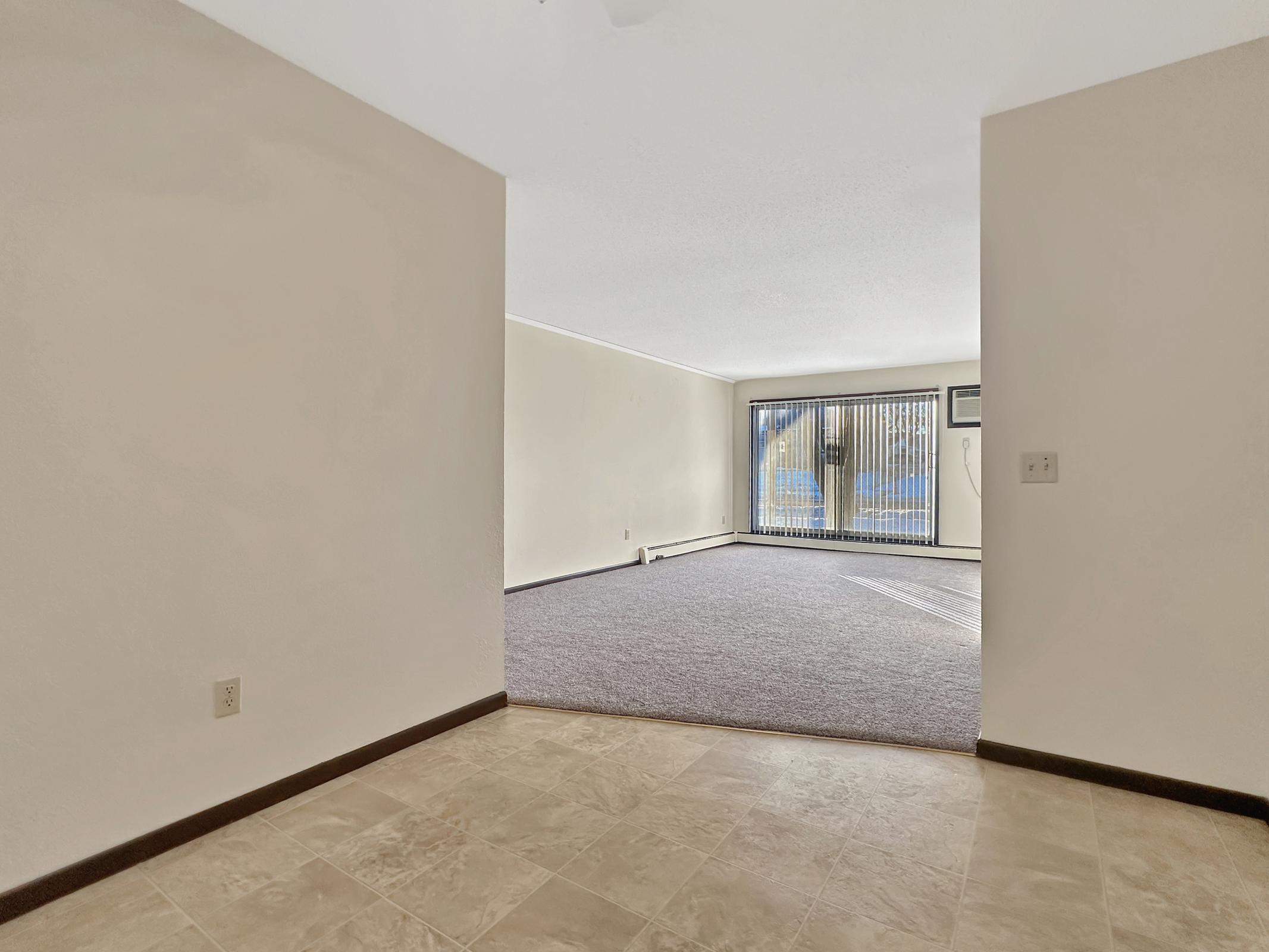 A view from a hallway into a bright, empty living room with a large window featuring vertical blinds. The flooring transitions from tiled to carpeted, with a wall-mounted air conditioning unit visible in the room. The walls are painted a neutral color, creating a spacious and airy atmosphere.