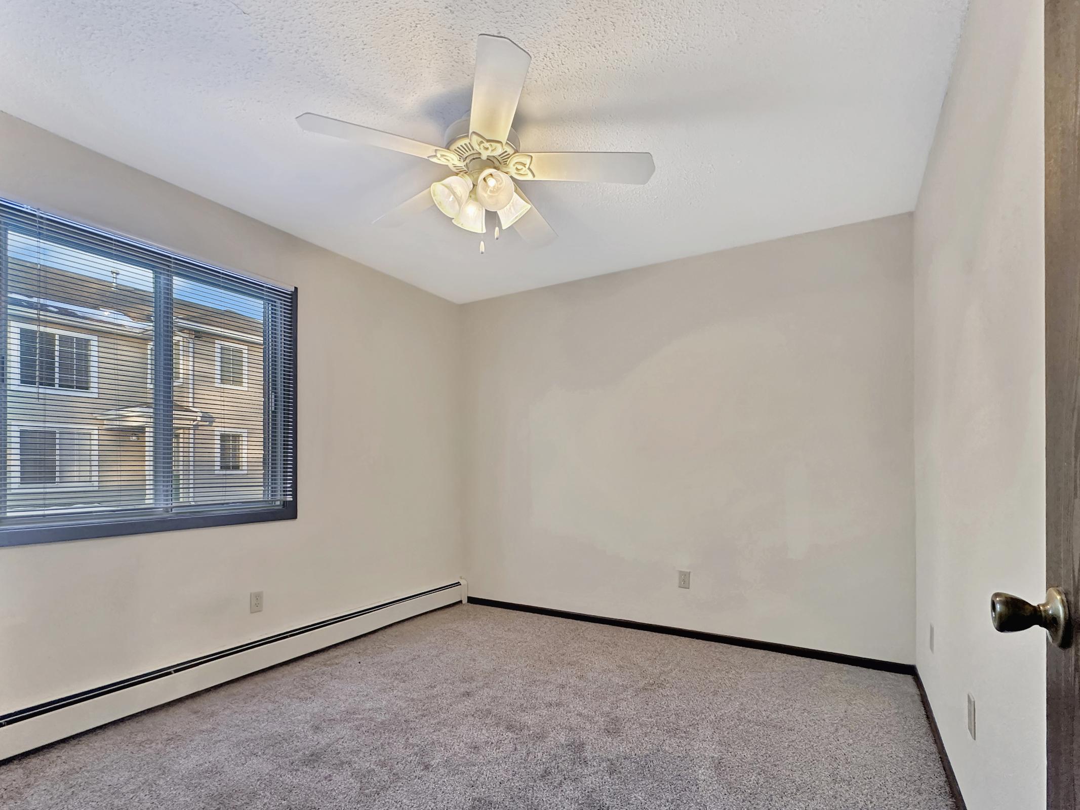 Empty room with light beige walls and gray carpet. A ceiling fan with four lights is mounted on the ceiling. There's a large window on one wall, allowing natural light to enter, with a view of neighboring buildings outside. The room has a simple, clean appearance.