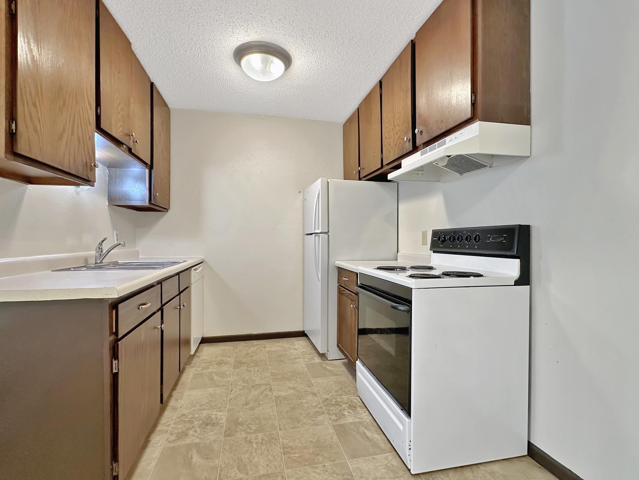A simple kitchen featuring wooden cabinets, a white stove, a refrigerator, and a double sink. The countertops are light-colored, and the flooring is tiled in a neutral tone. The walls are painted light, creating a spacious and clean look. A ceiling light provides illumination.