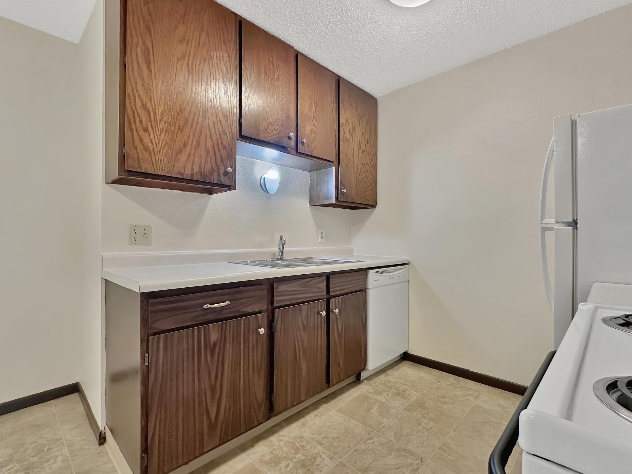 A compact kitchen featuring wooden cabinets, a sink with a faucet, and a dishwasher. The countertop is white, and there is a white refrigerator and stove in the image. The walls are light-colored, and the flooring is tiled. The space is well-lit and organized, emphasizing a functional design.