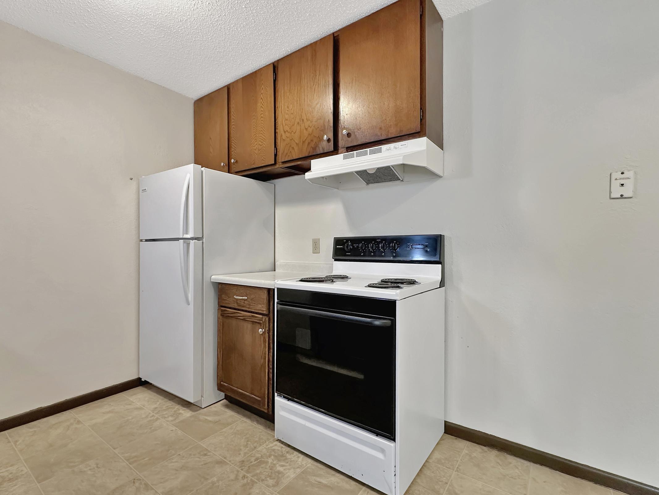A small kitchen featuring a white refrigerator, a black stove and oven, and a range hood. The cabinetry above is dark wood, and the countertops are light-colored. The floor has beige tiles, and the walls are a light gray shade.