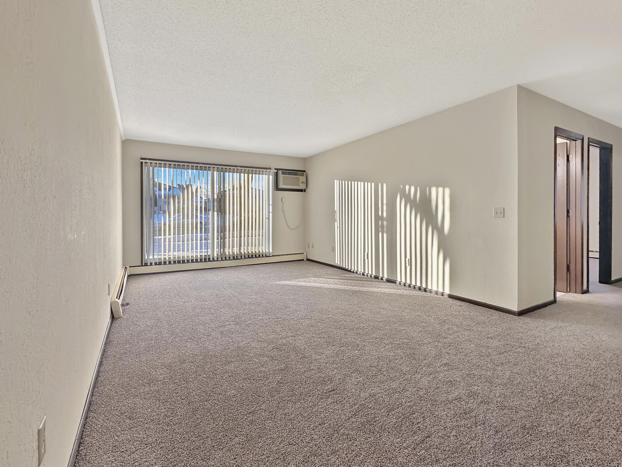 Bright, empty living room with light beige walls and carpet. Large window with vertical blinds allows natural light to fill the space. An air conditioning unit is visible on the left wall. The room features a clean, minimalist design with a door leading to another area on the right.