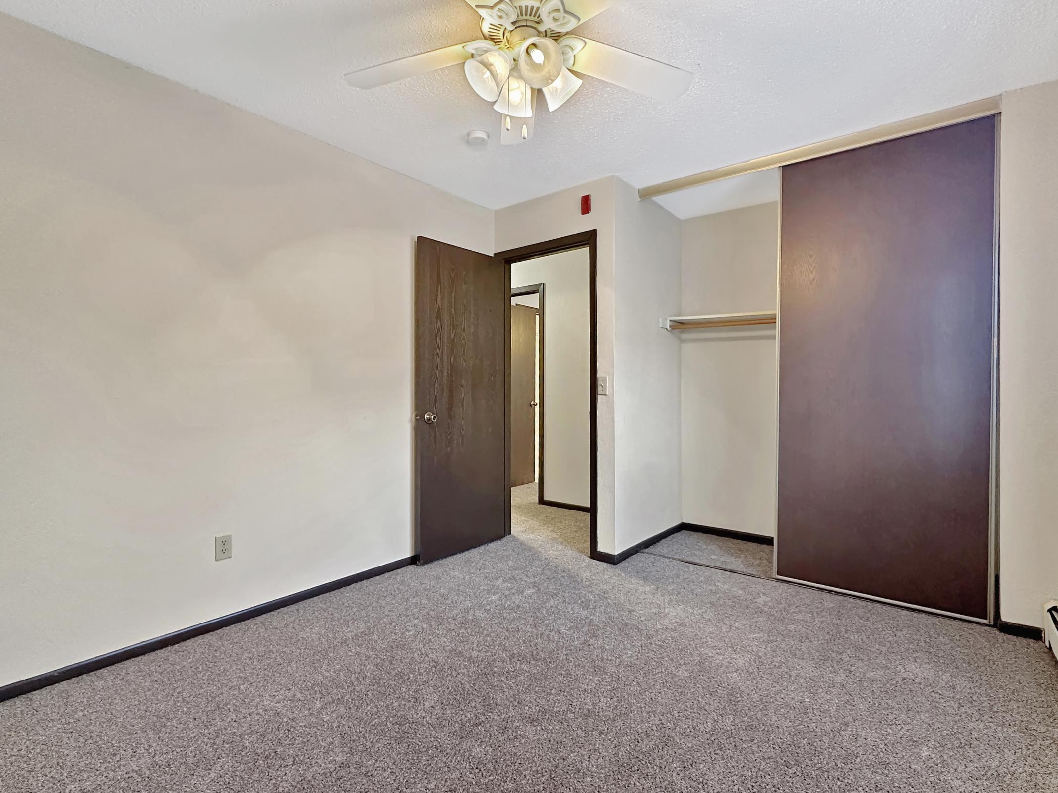 A well-lit, empty room with light-colored walls, a carpeted floor, and a ceiling fan. There is a door on one side leading to another area, and a closet space with sliding doors on the opposite wall. The overall atmosphere is clean and spacious, suitable for various uses.