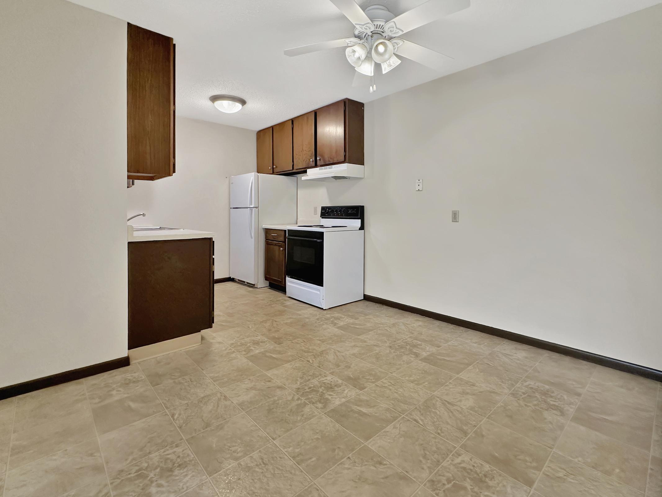 A clean, modern kitchen space with brown cabinetry, a white refrigerator, and a black stove. The floor is tiled in light beige, and there's a ceiling fan. The walls are painted a light color, creating a bright atmosphere.