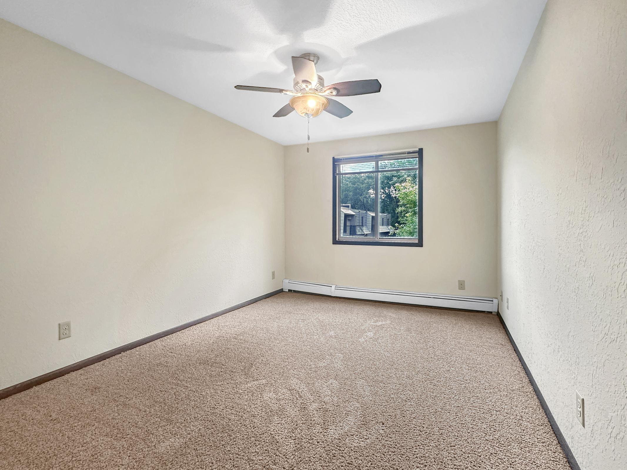 Empty room with light beige walls and a ceiling fan. The floor is covered in light brown carpet. A single window allows natural light to enter, revealing greenery outside. The space appears clean and uncluttered, suggesting it's ready for new furnishings or decor.