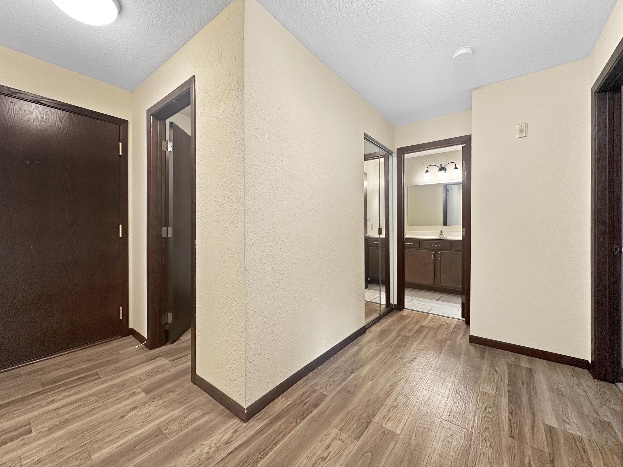 A well-lit hallway featuring beige walls, dark wood trim, and a laminate floor. On one side, there’s a mirror and a doorway leading to a bathroom with dark cabinetry. To the left, the entrance door is visible. The layout suggests a modern apartment or condo design.