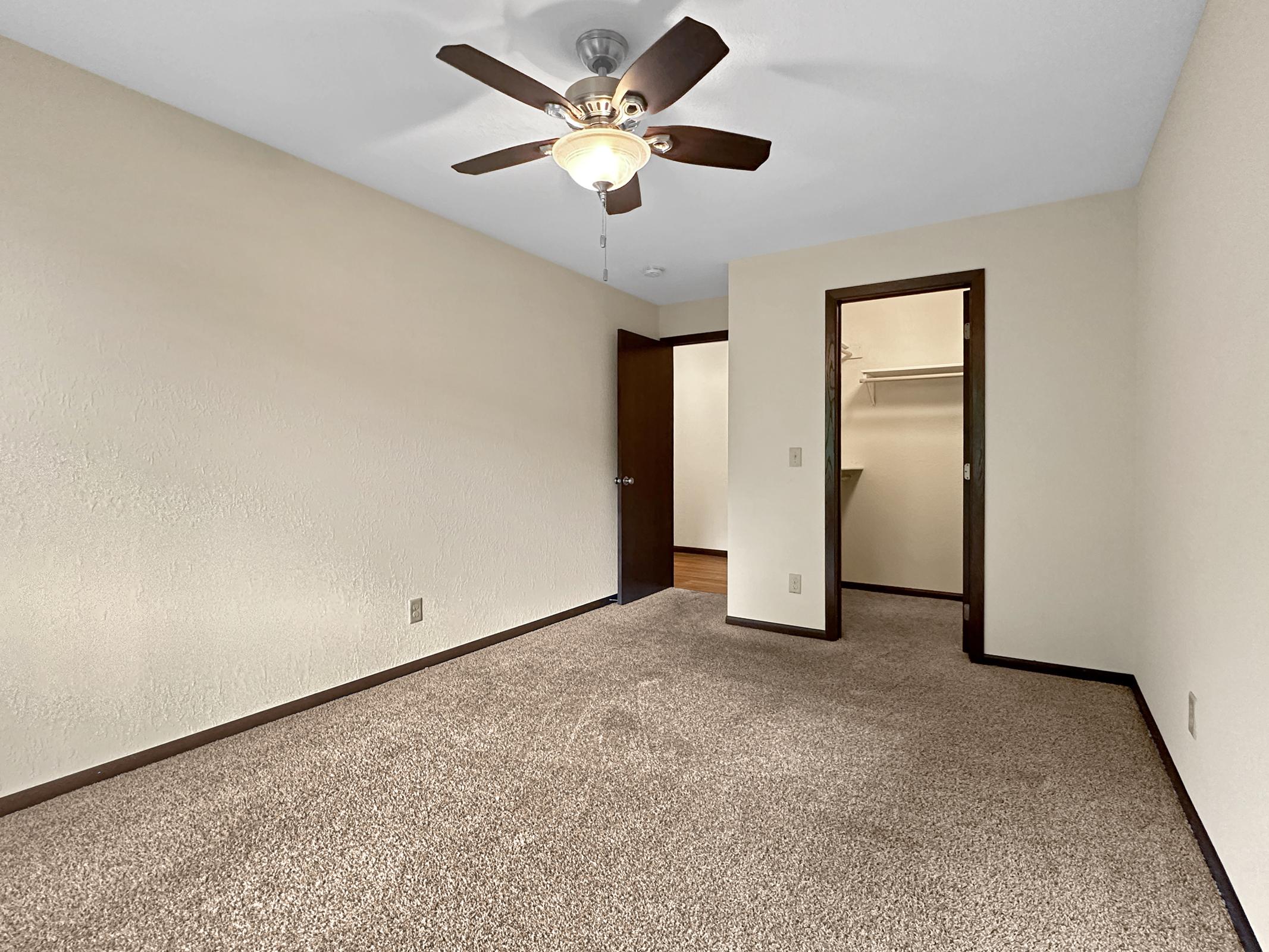 A spacious, empty room featuring beige walls and light brown carpet. A ceiling fan with wooden blades hangs centrally. There are two doorways: one leading to a closet and another to an adjoining space. The room is well-lit, showcasing its potential as a living or bedroom area.