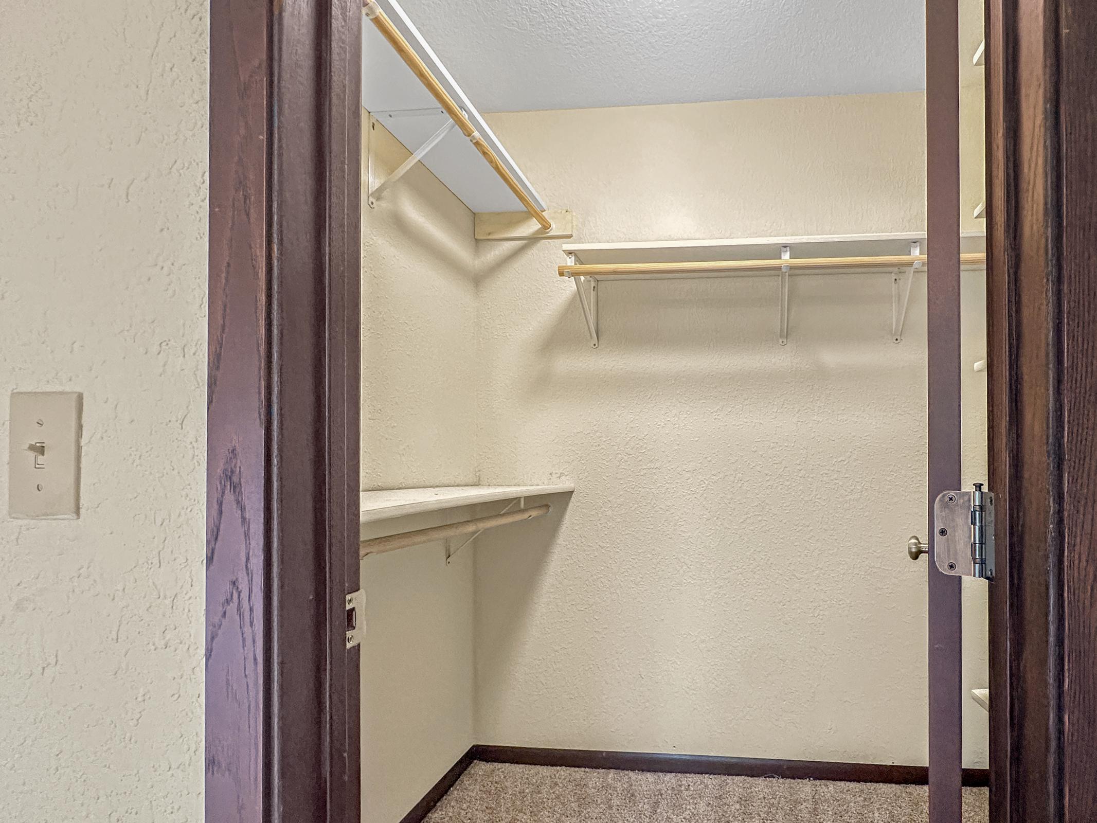 Interior of a spacious empty closet featuring beige walls, a carpeted floor, and wooden door frames. The closet has shelves and hanging rods for storage but contains no items, showcasing a clean and organized space.